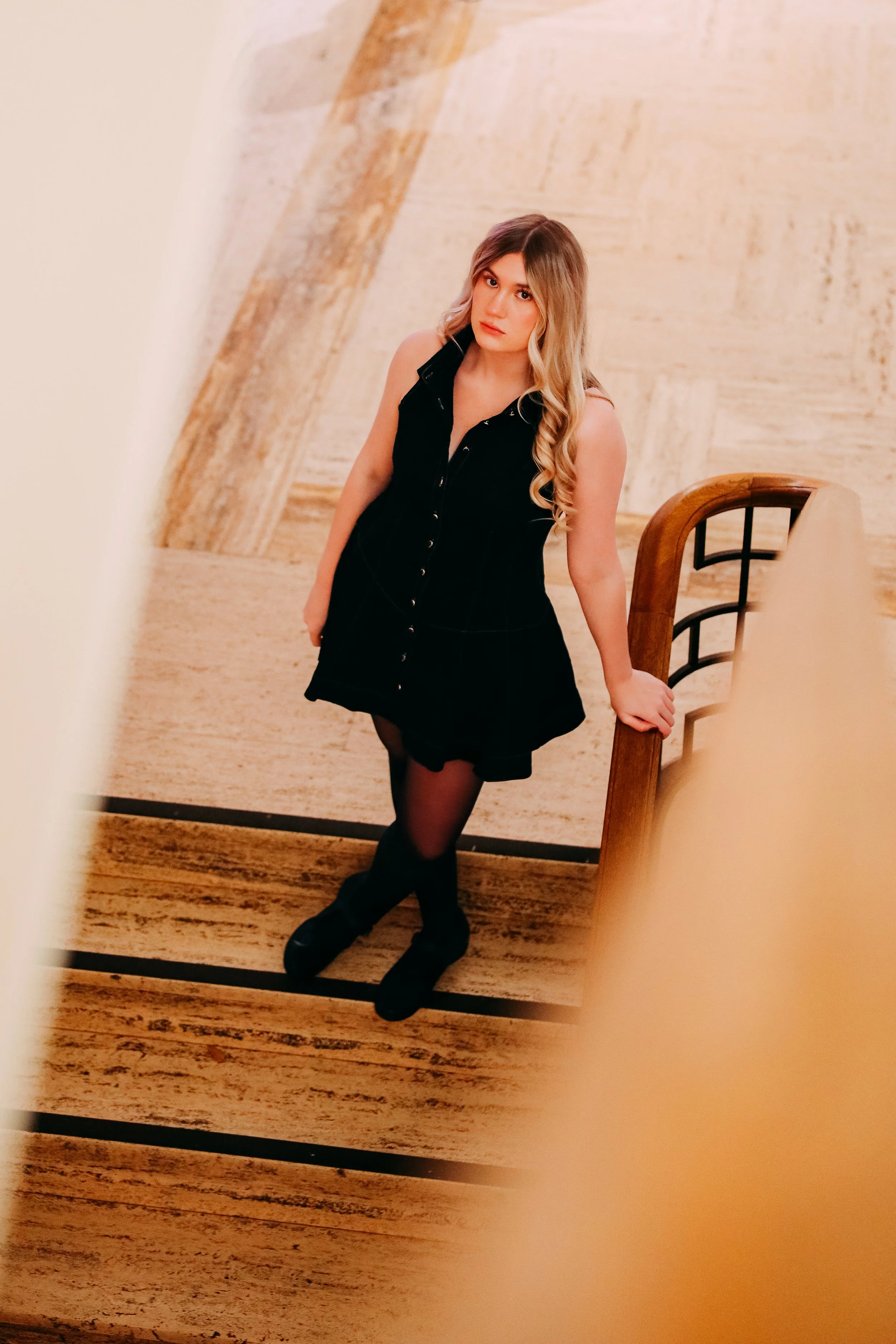 A young woman with long blonde hair, wearing a black sleeveless dress, black tights, and black shoes, posing indoors while holding onto a wooden railing.