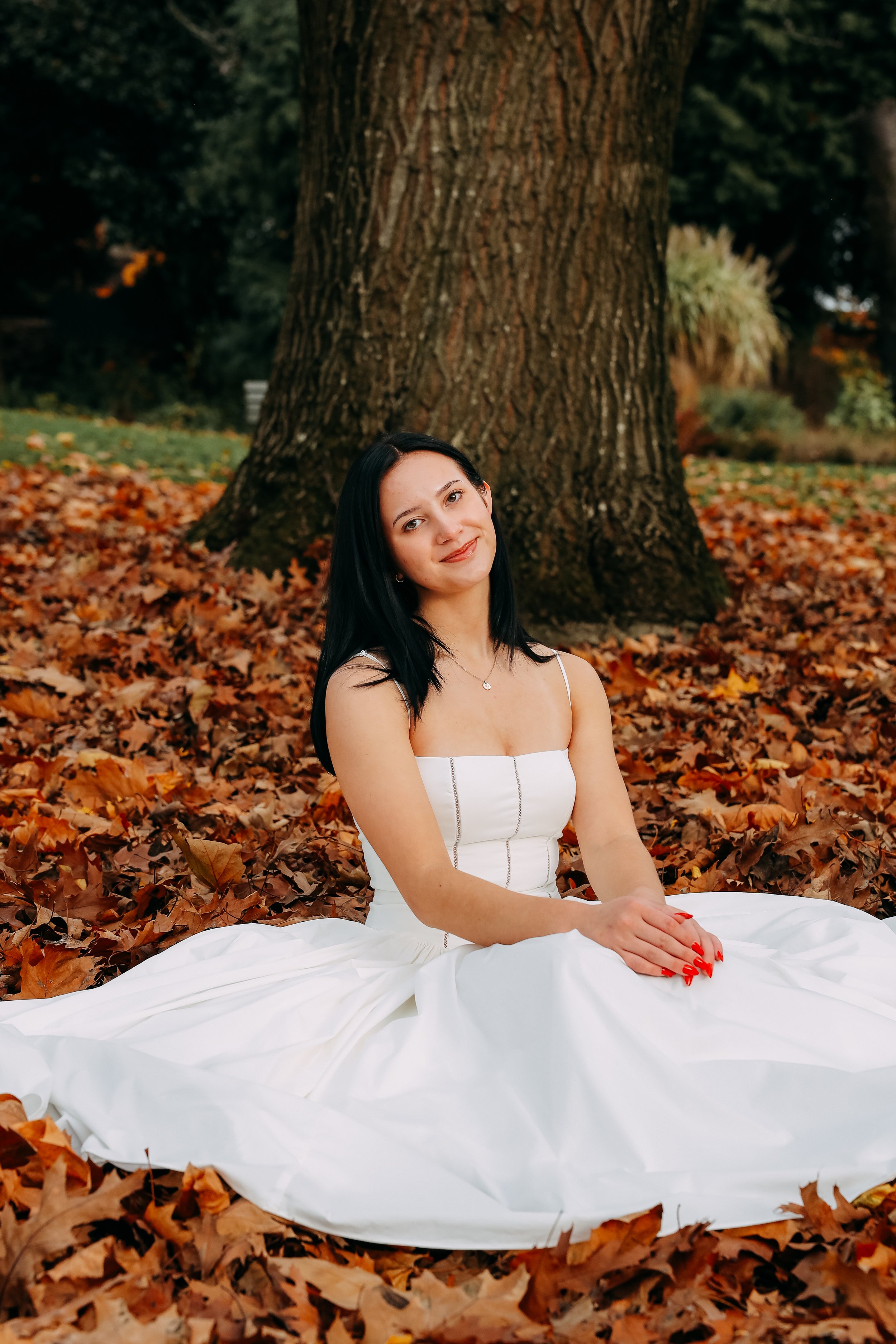 A woman in a white dress sitting on fallen autumn leaves in front of a large tree in a park.
