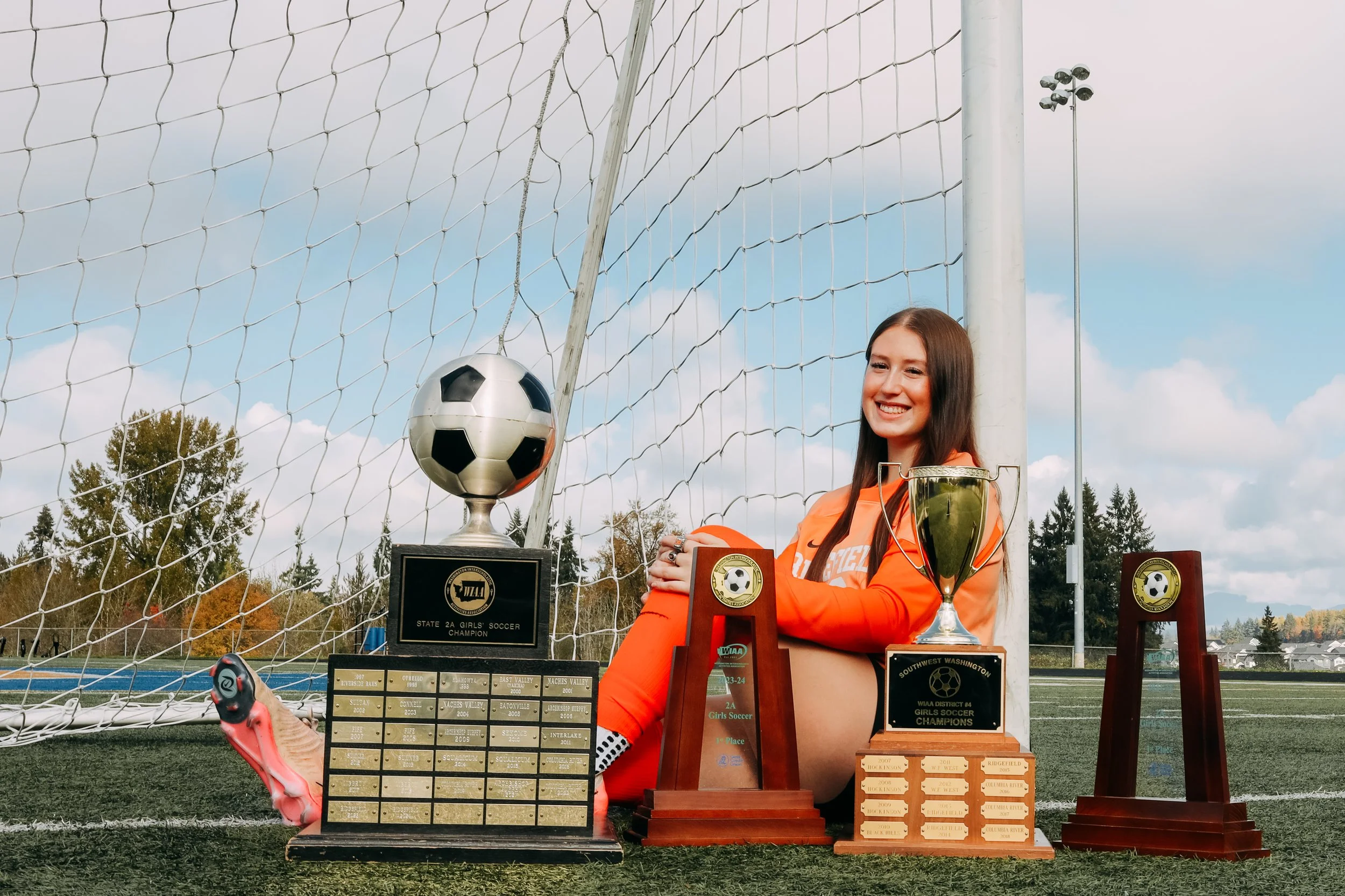 A young female soccer player in an orange uniform sitting on a soccer field surrounded by trophies and awards, with a soccer net and trees in the background.