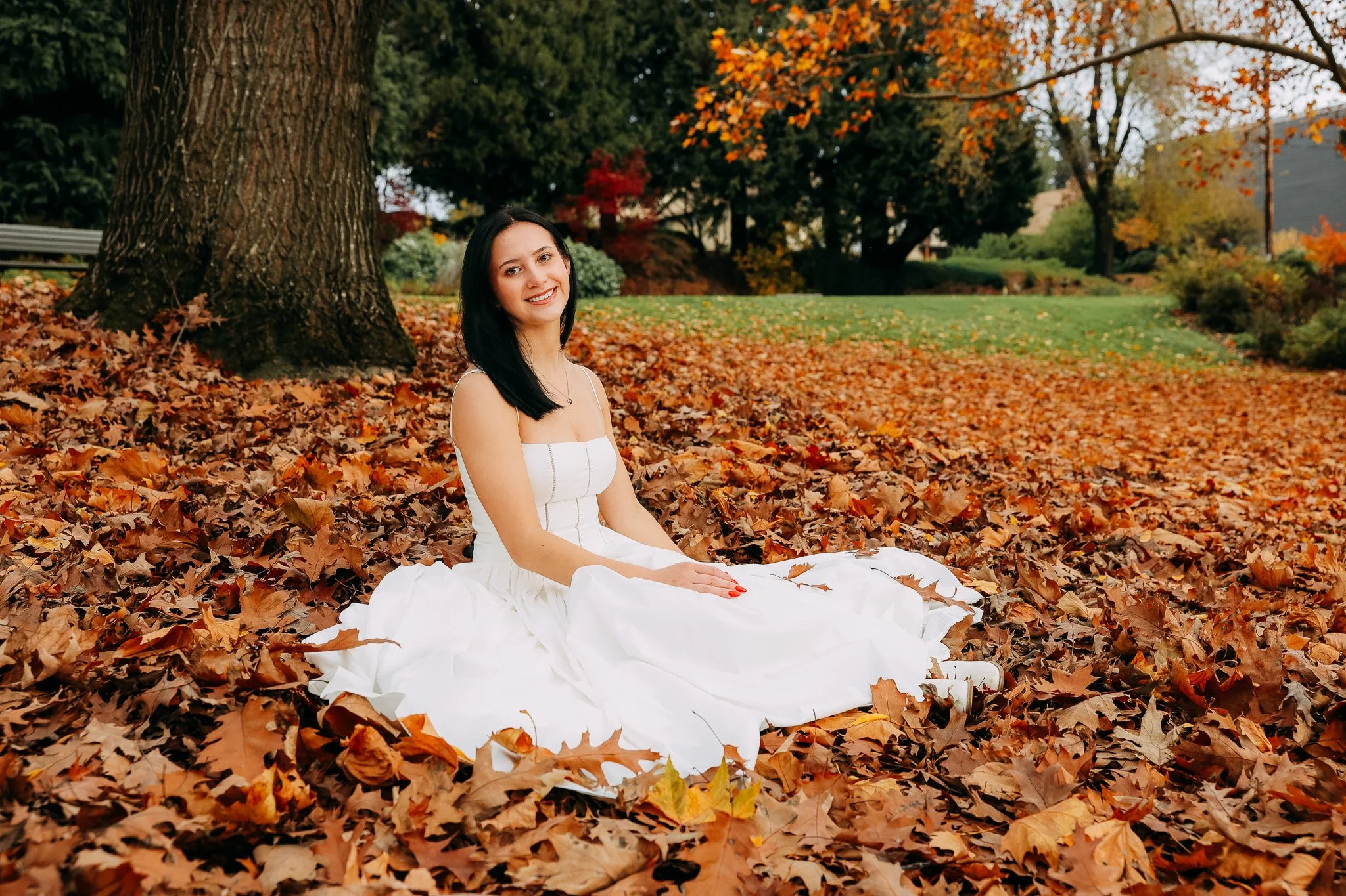 A young woman in a white dress sitting on fallen autumn leaves in a park with trees and colorful fall foliage.