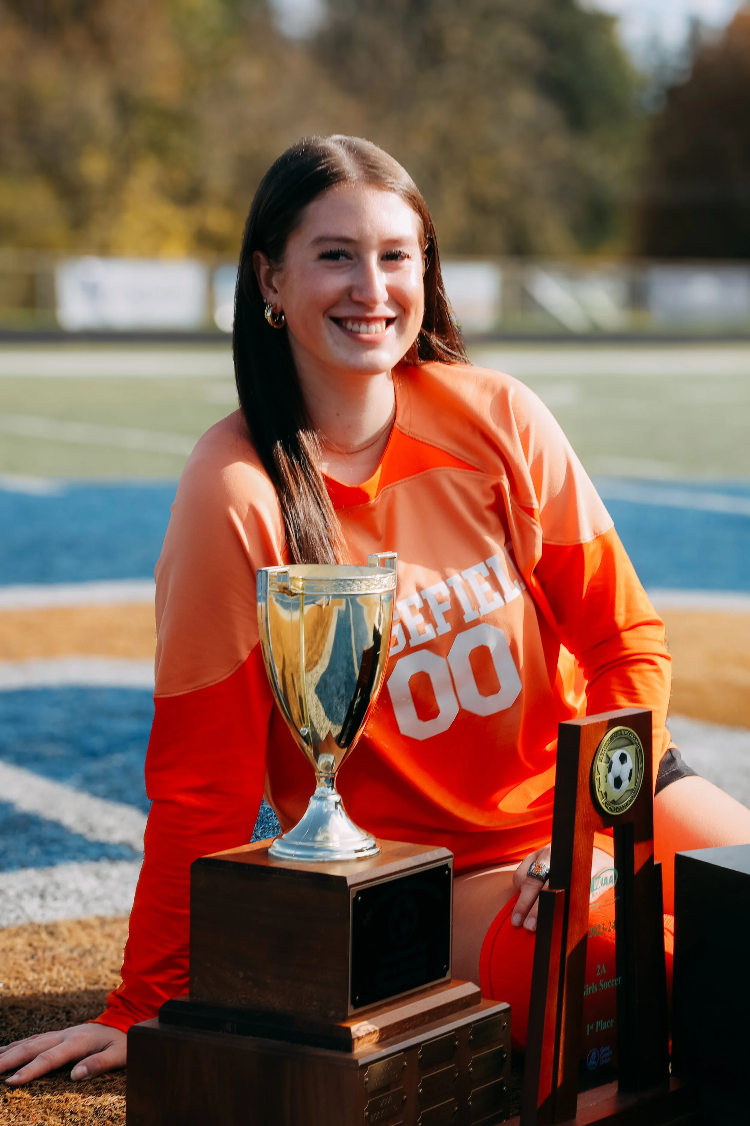 A young woman in an orange soccer jersey with the number 00, sitting on a soccer field with trophies in front of her, smiling at the camera after a victory.