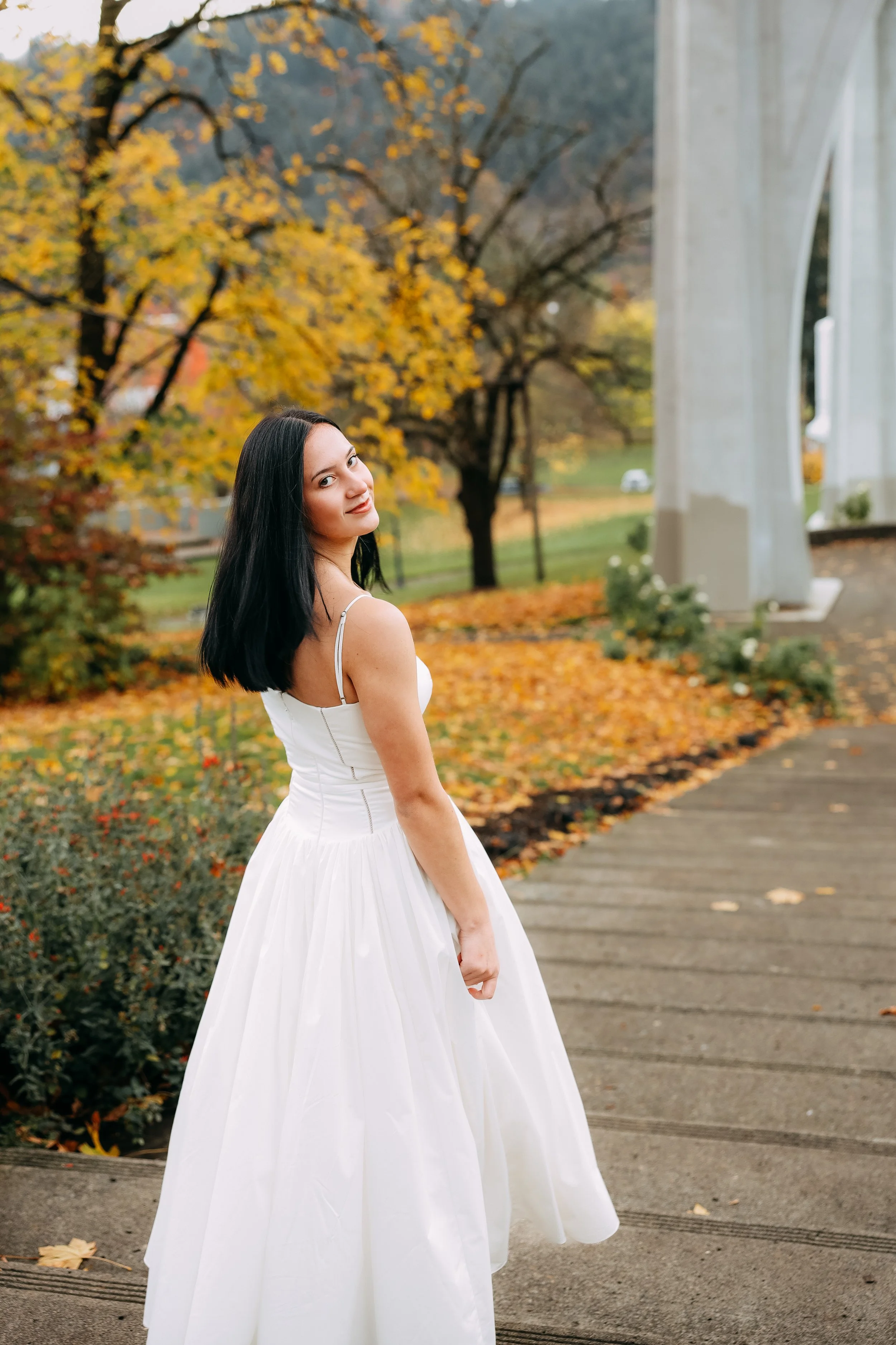 A woman in a white dress standing on a sidewalk in an autumn park, with yellow and orange leaves on trees and the ground, near a white bridge.