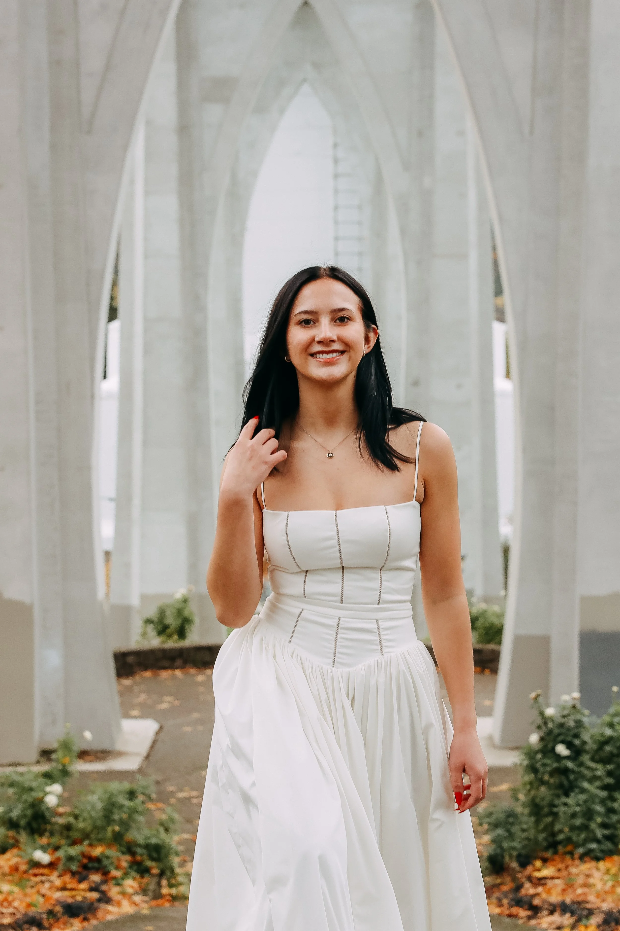 A young woman in a white dress standing outdoors in front of a modern concrete sculpture with arches, smiling and touching her hair.