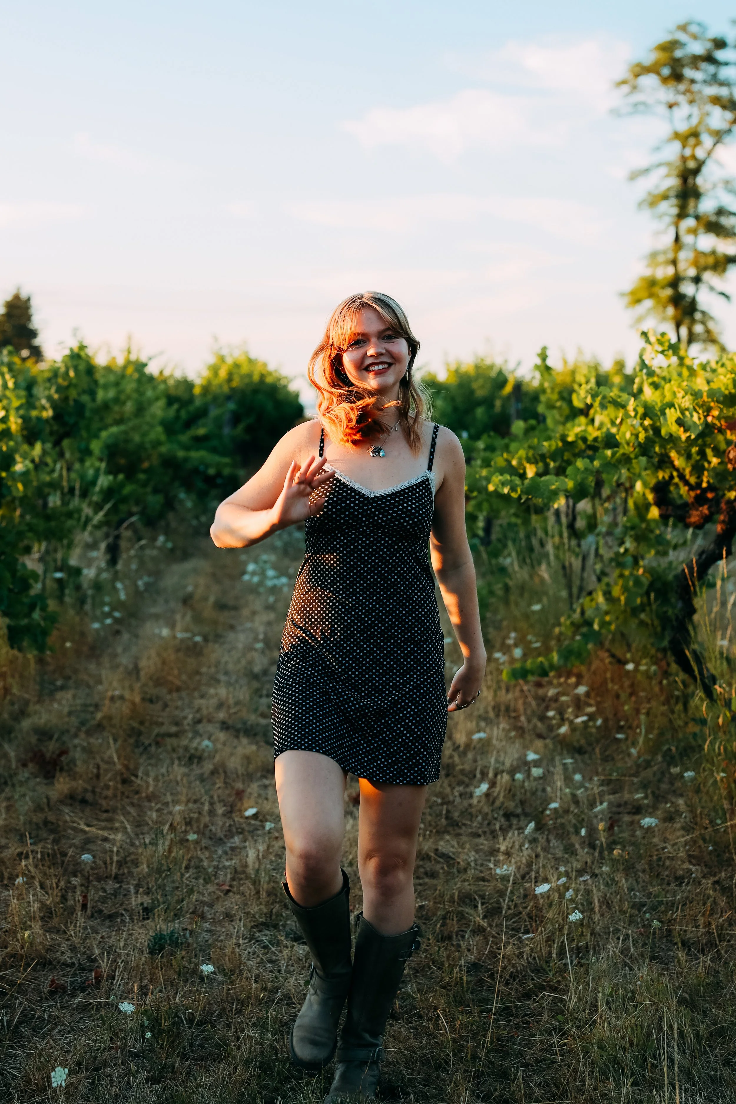 A young woman with shoulder-length brown hair, wearing a black dress with white polka dots and black boots, walking through a vineyard during sunset and smiling.