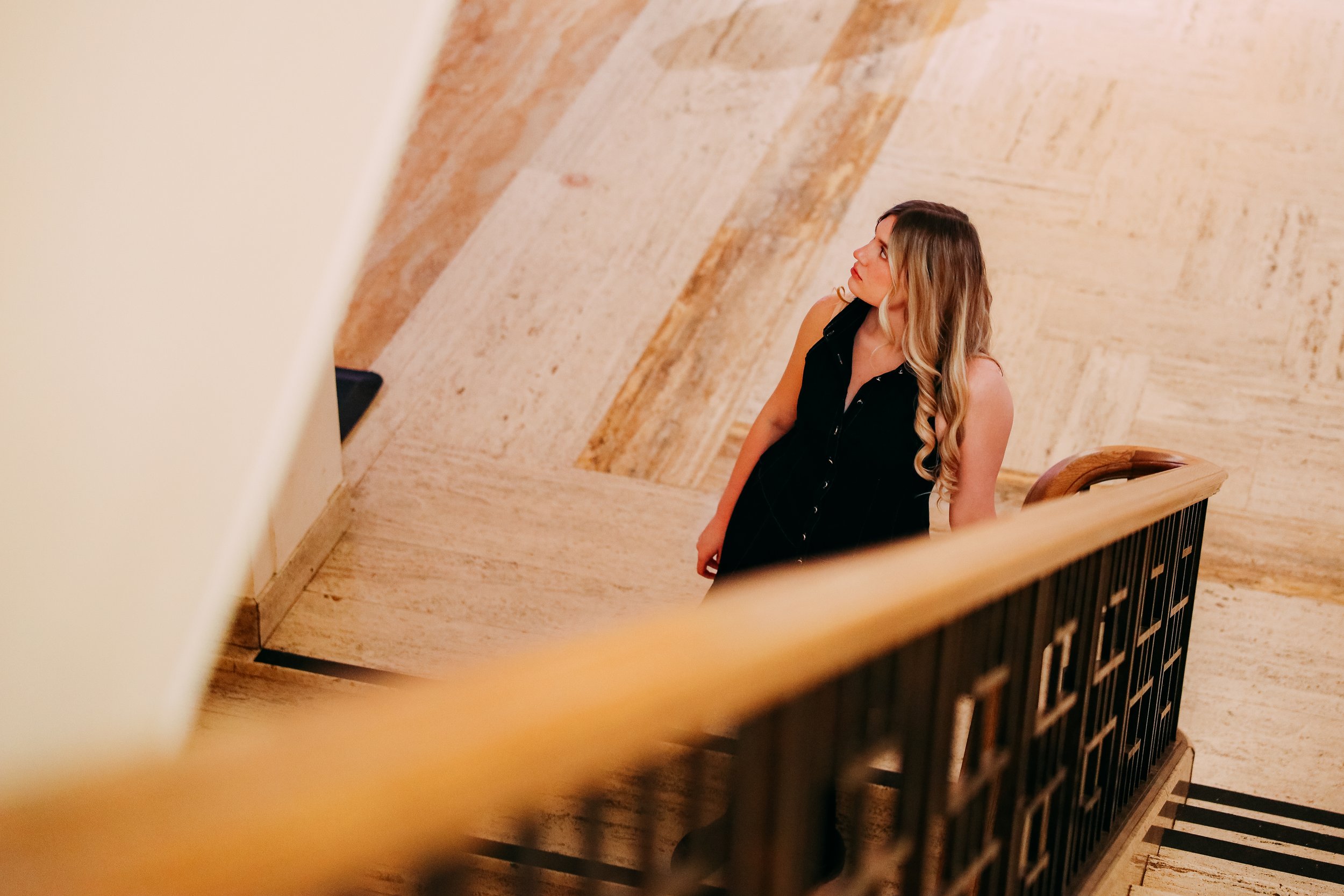 Woman with long blonde hair, wearing a sleeveless black dress, standing on a staircase, looking to her right.