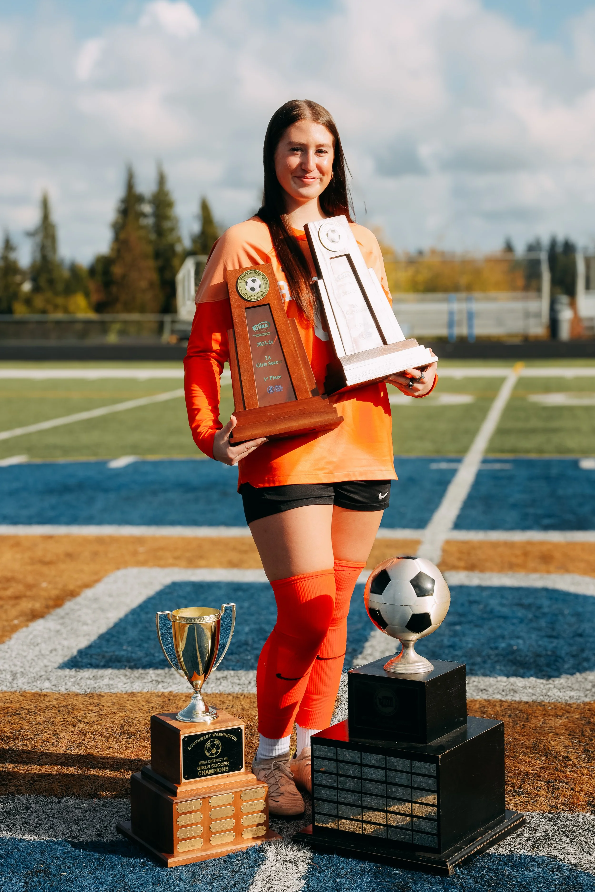 A young female soccer player in an orange and black uniform holding two trophies, standing on a soccer field with additional trophies at her feet.