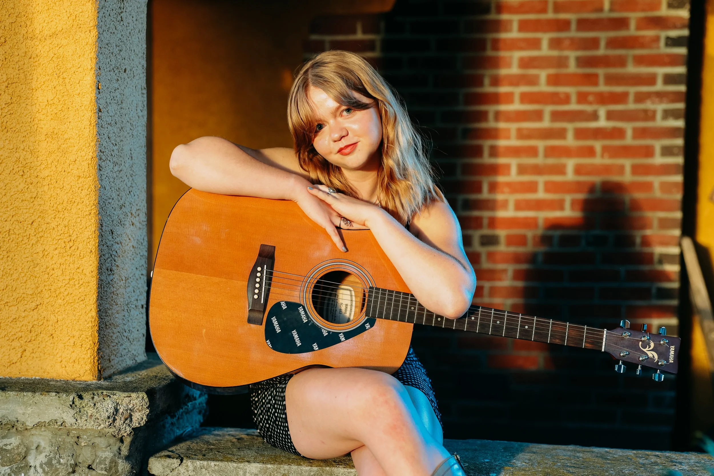 A young woman with blonde hair sitting outdoors against a brick wall, holding an acoustic guitar and looking at the camera.