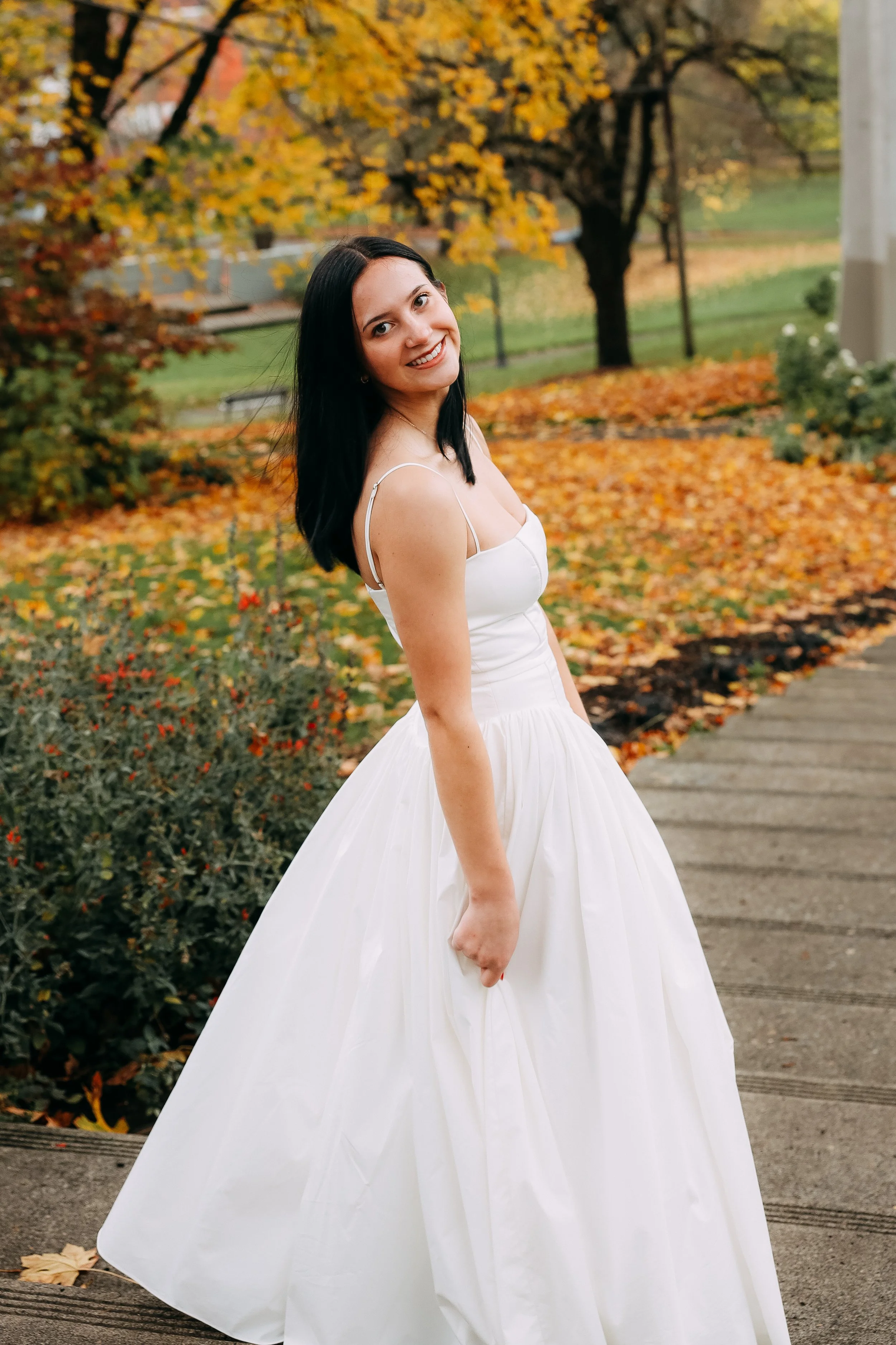 A young woman in a white dress standing outdoors on a sidewalk with autumn leaves and trees in the background.