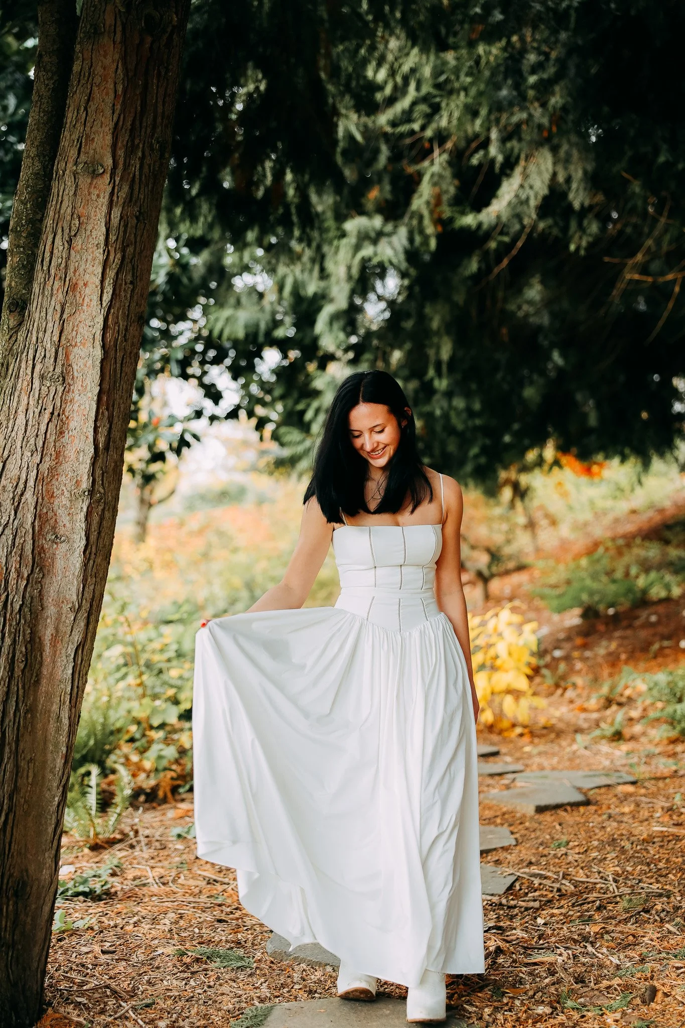 A woman in a white dress walking on a stone pathway in a wooded outdoor setting, smiling and looking down at her dress.