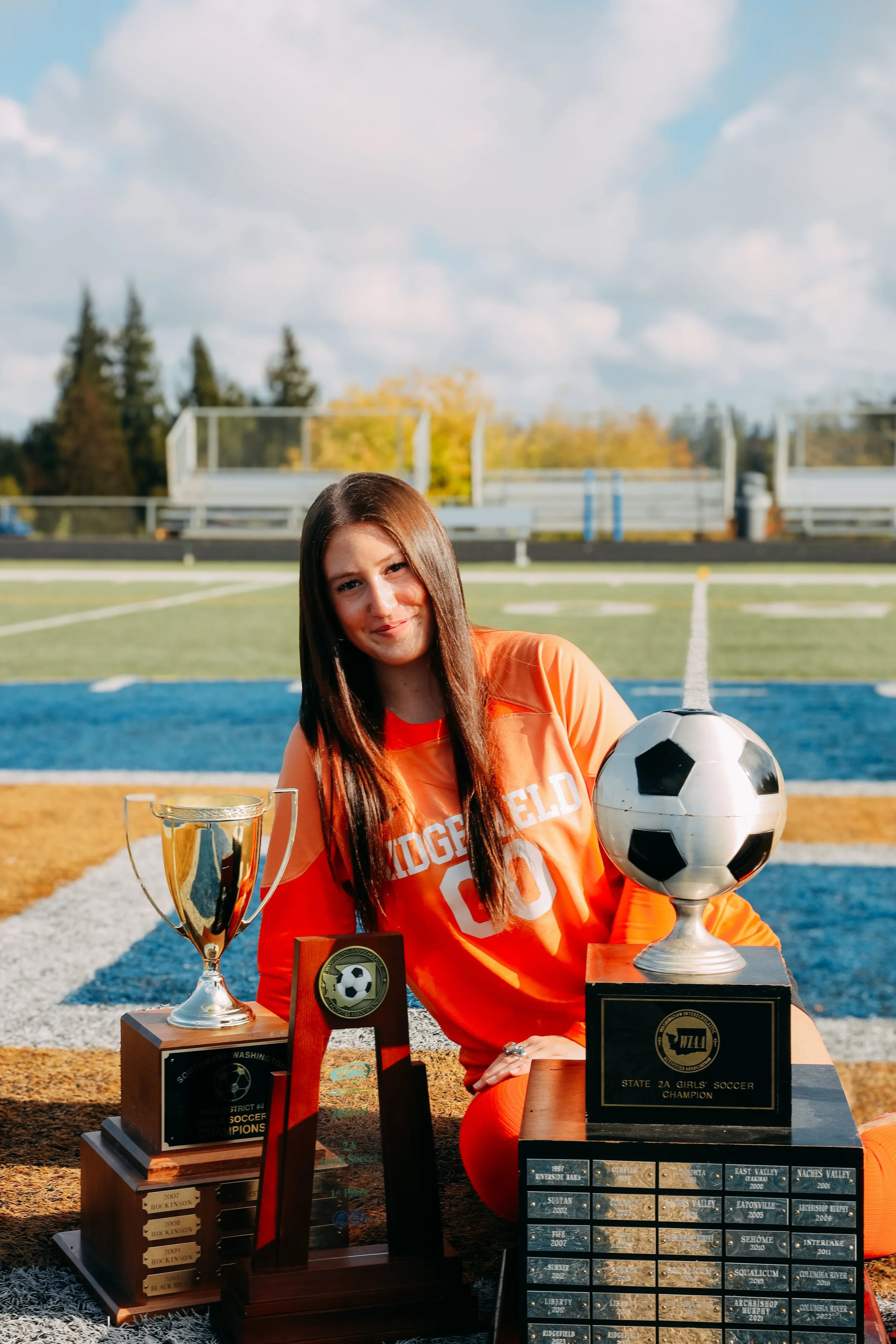 Young female soccer player in orange uniform sitting on soccer field with medals and trophies.