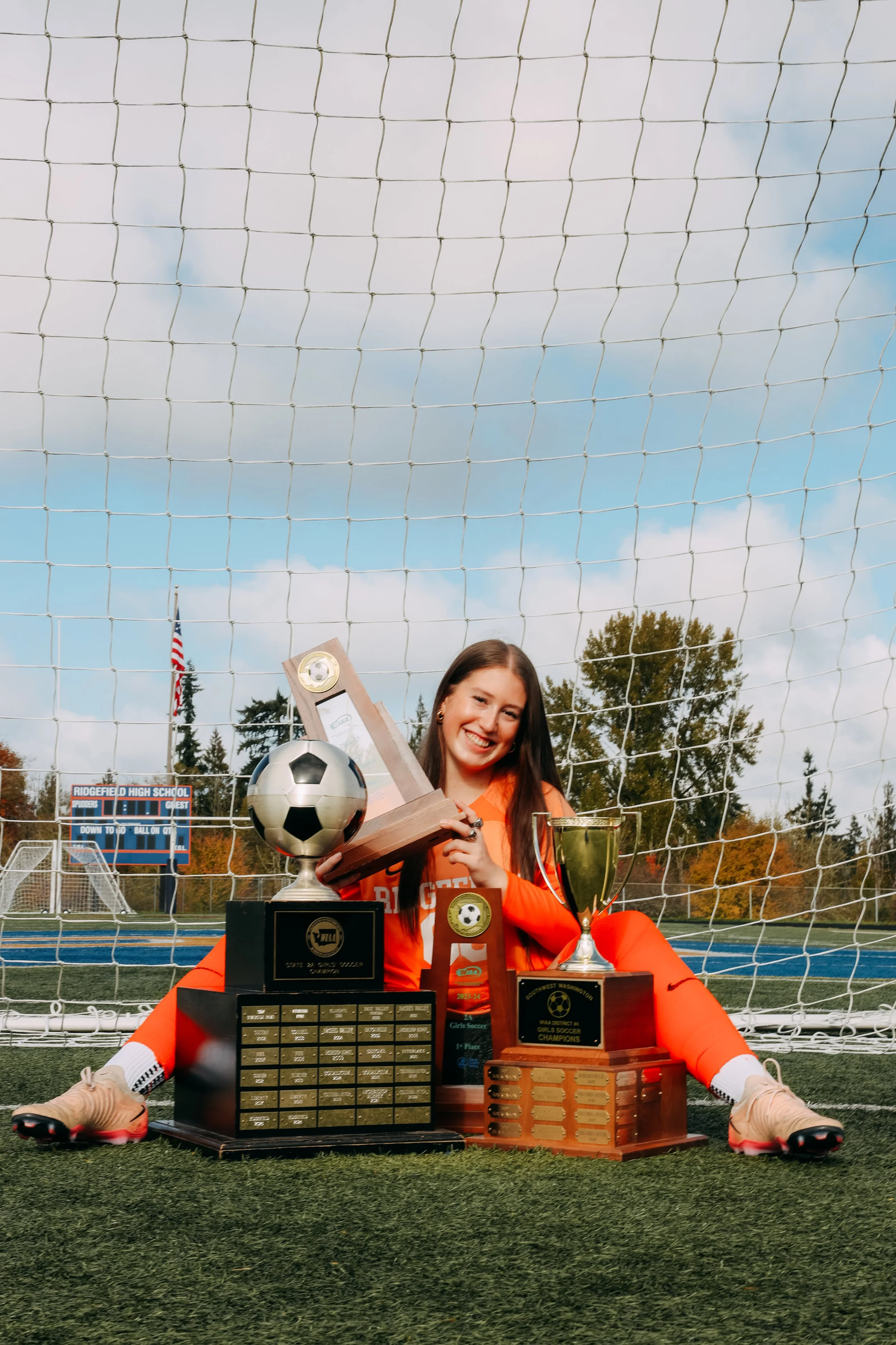A young woman in soccer gear sitting on a soccer field surrounded by soccer trophies and awards.