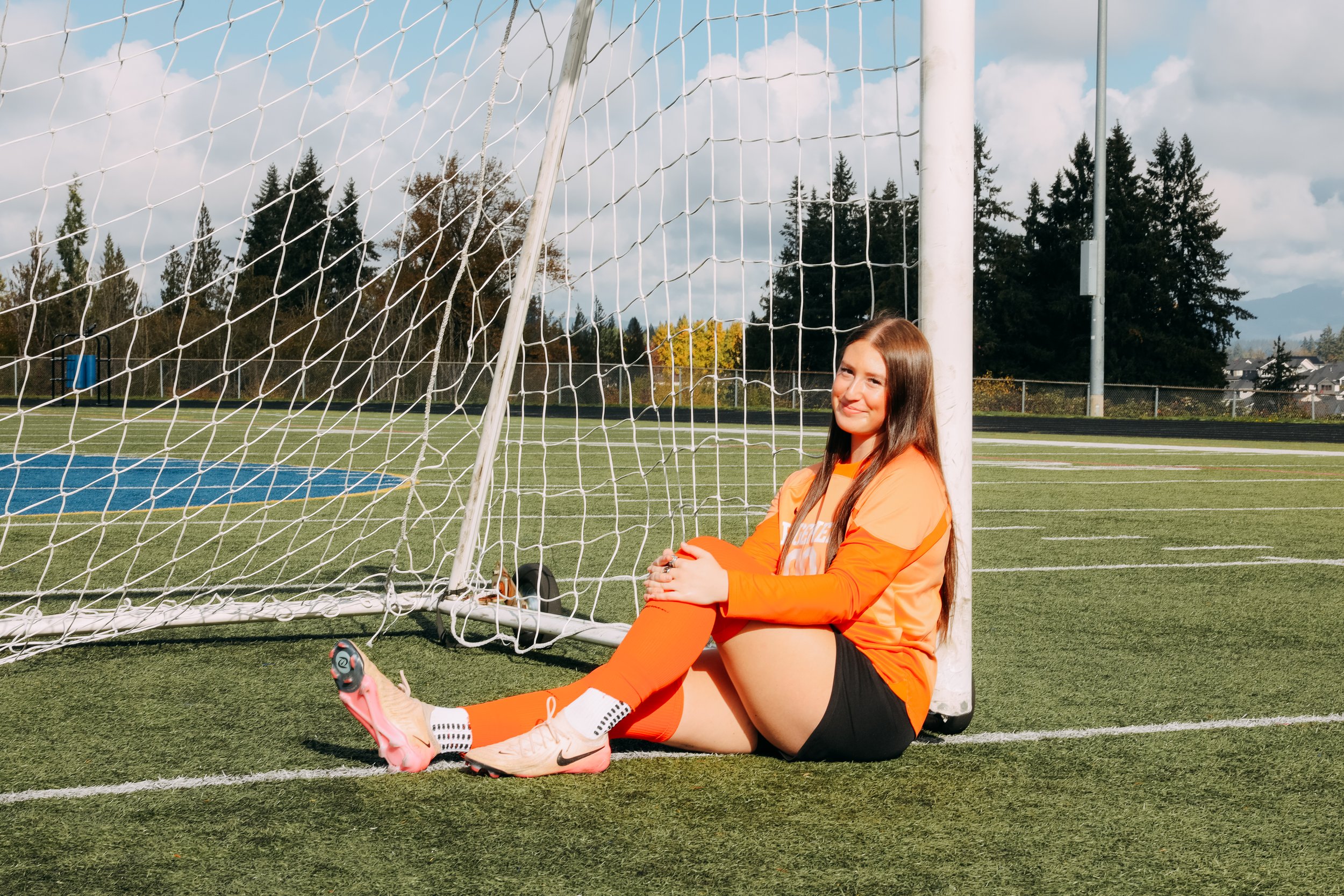 A young woman wearing an orange soccer jersey and black shorts sitting on the field next to a soccer goalpost, smiling at the camera with trees and a blue sky in the background.