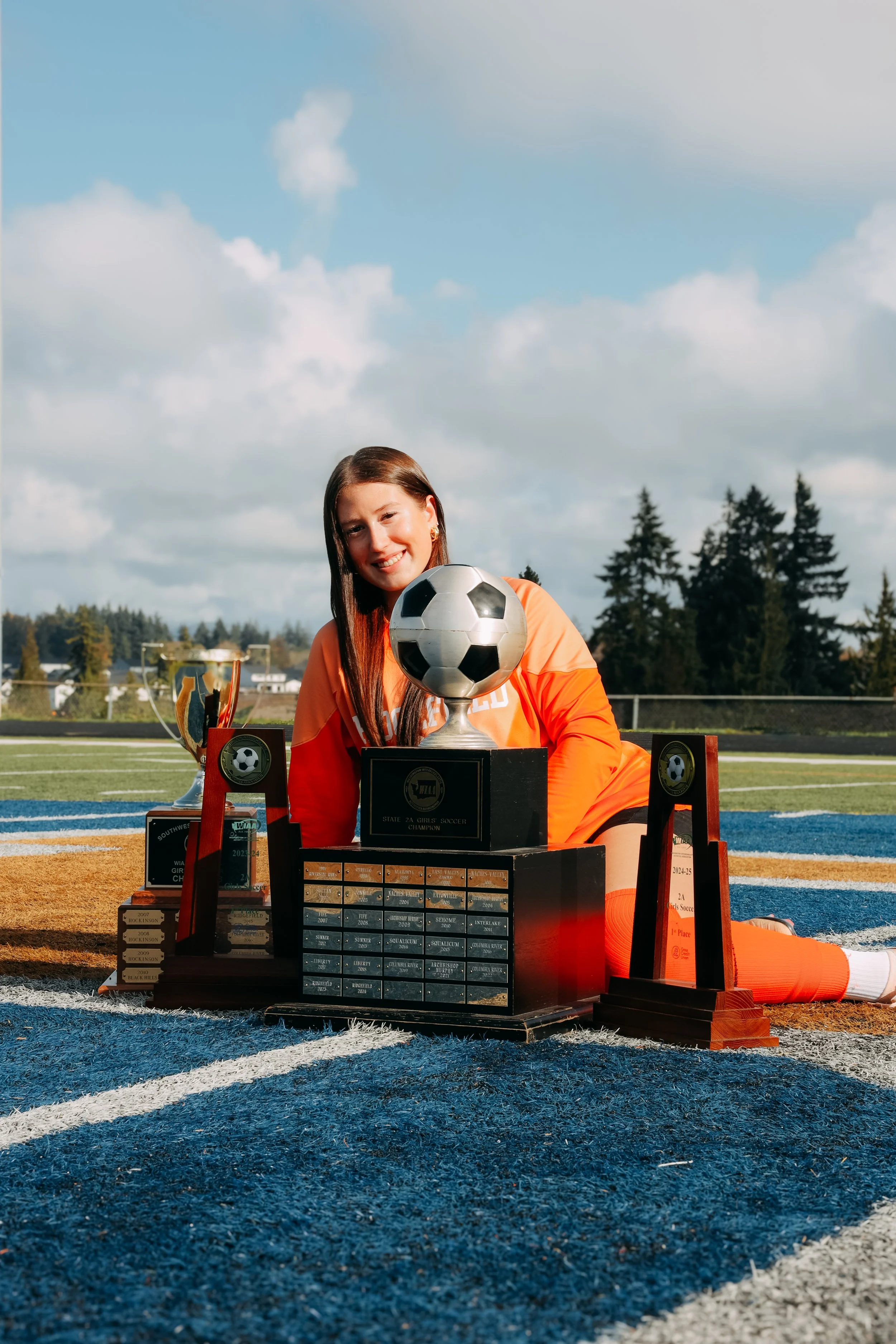 A young female soccer player in an orange uniform kneeling on a soccer field, smiling, surrounded by trophies and awards, with a large soccer ball trophy in front.