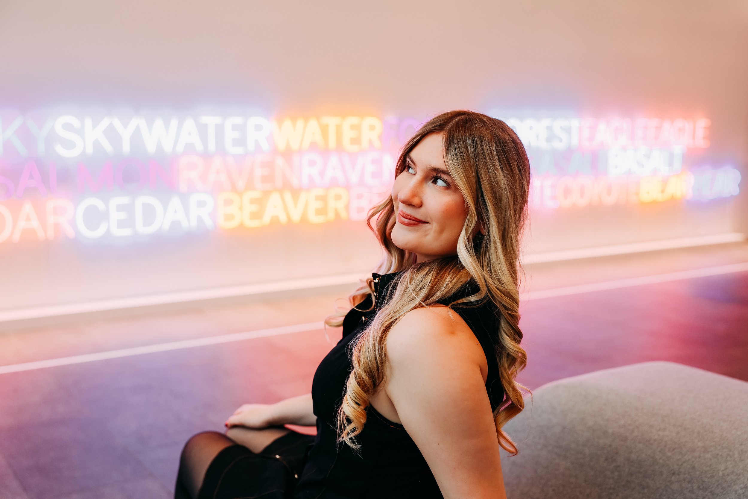 A young woman sitting on the floor with colorful neon signs in the background that display city names.