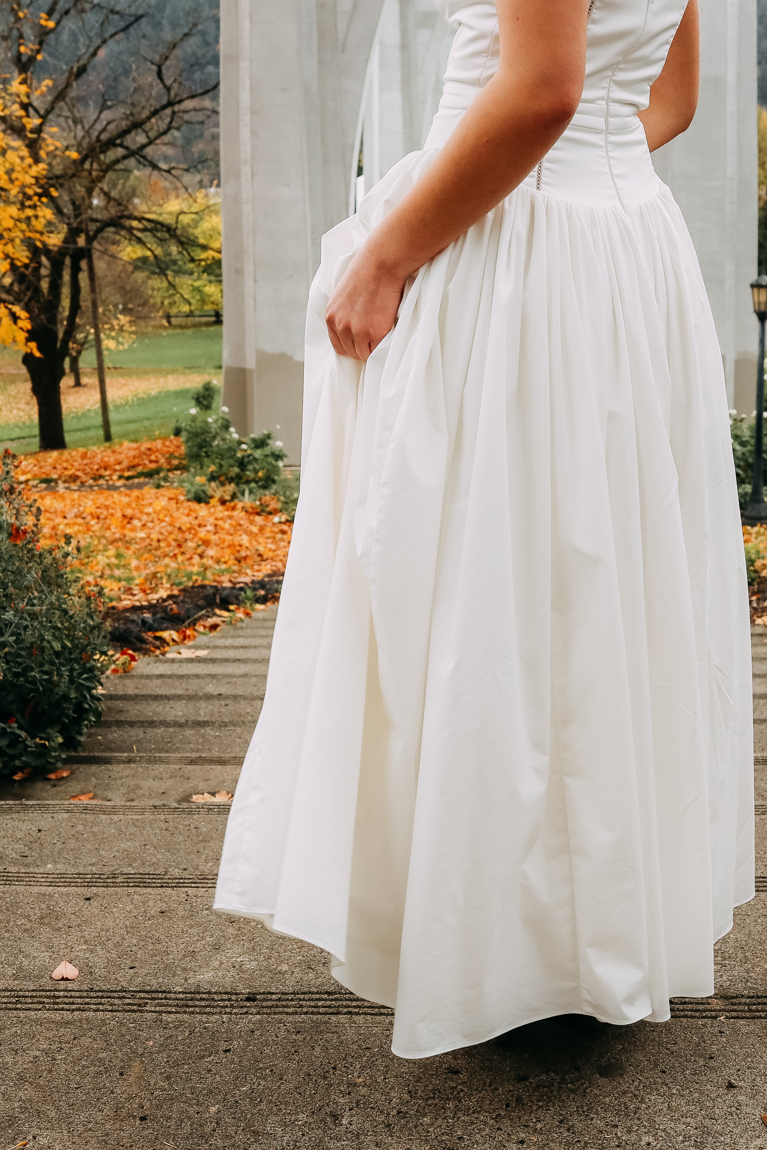 A woman in a white dress standing outdoors during fall, with autumn leaves on the ground and trees in the background.