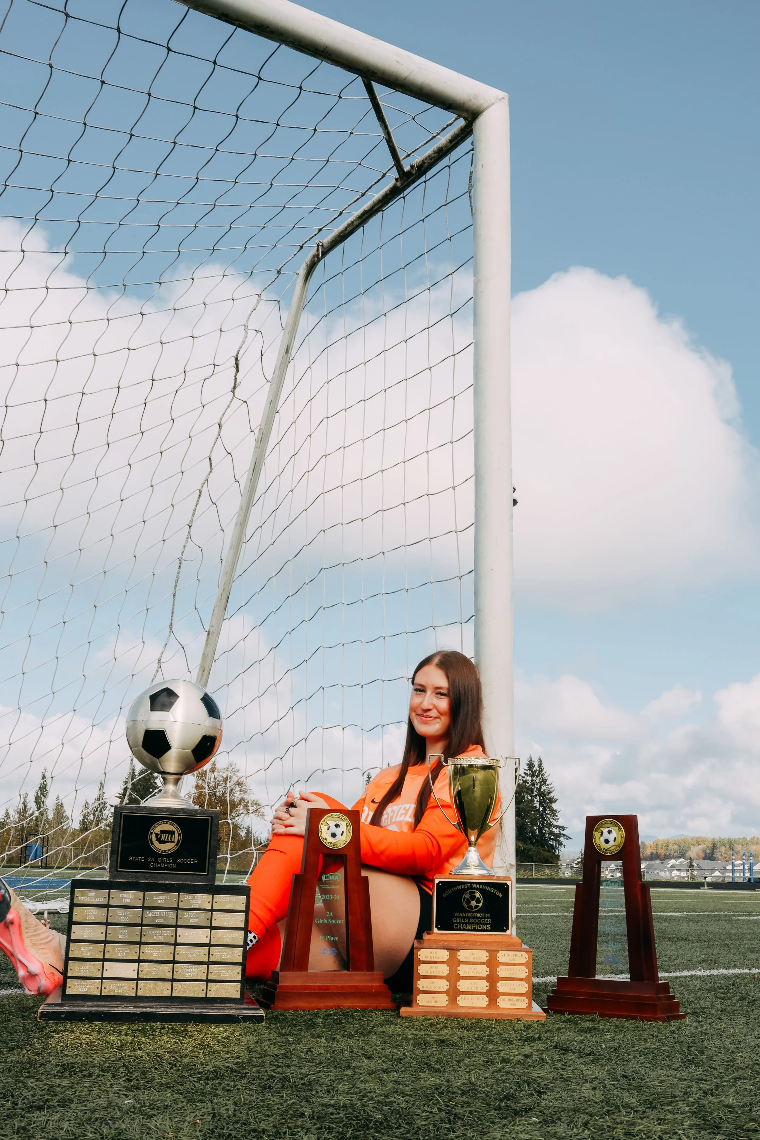 A young female soccer player in an orange uniform sitting on a soccer field with several trophies and awards around her, including a soccer ball trophy, a cup, and plaques, in front of a goalpost.