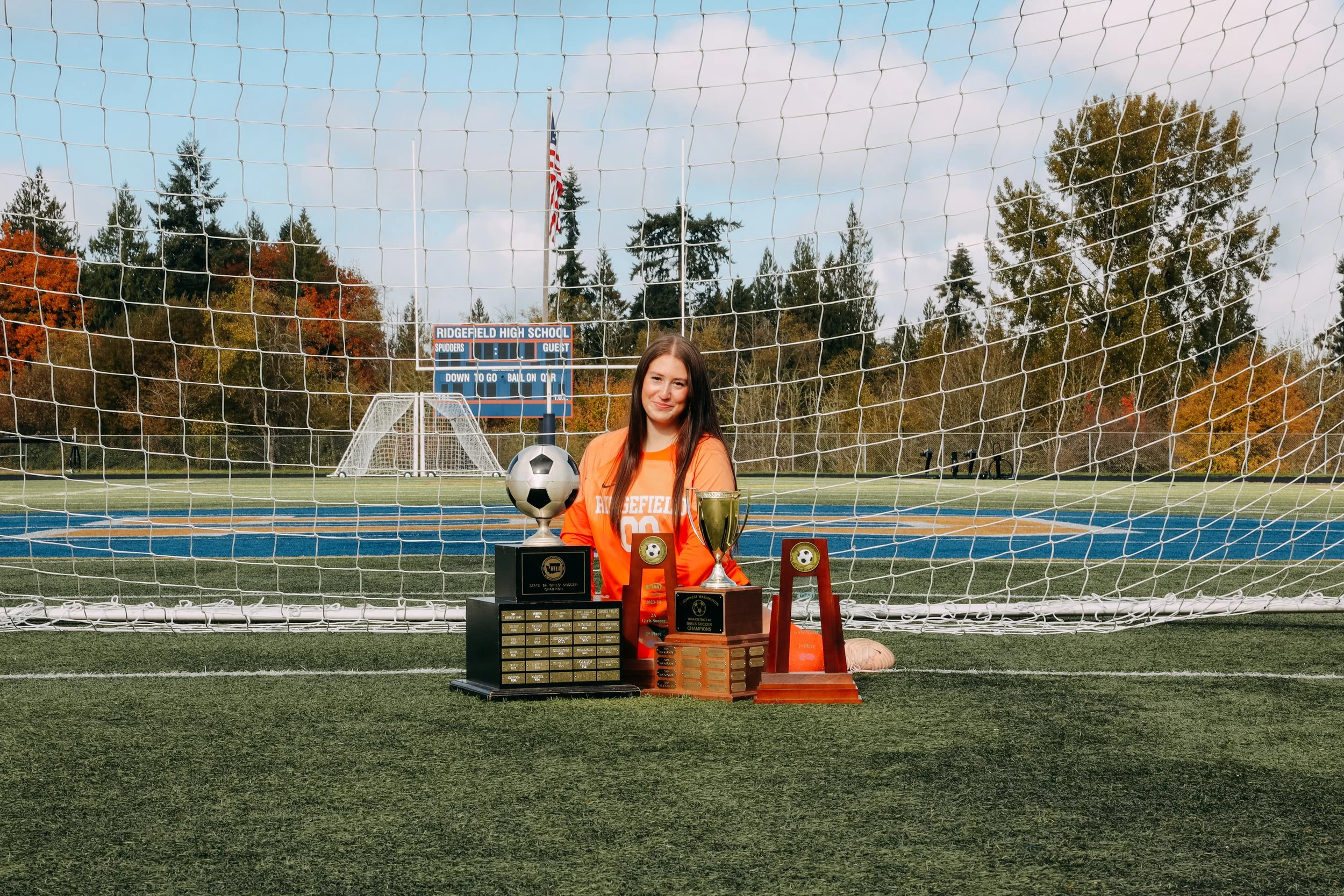 Young woman in orange soccer jersey sitting behind trophies on a soccer field, with a goalpost and scoreboard in the background.