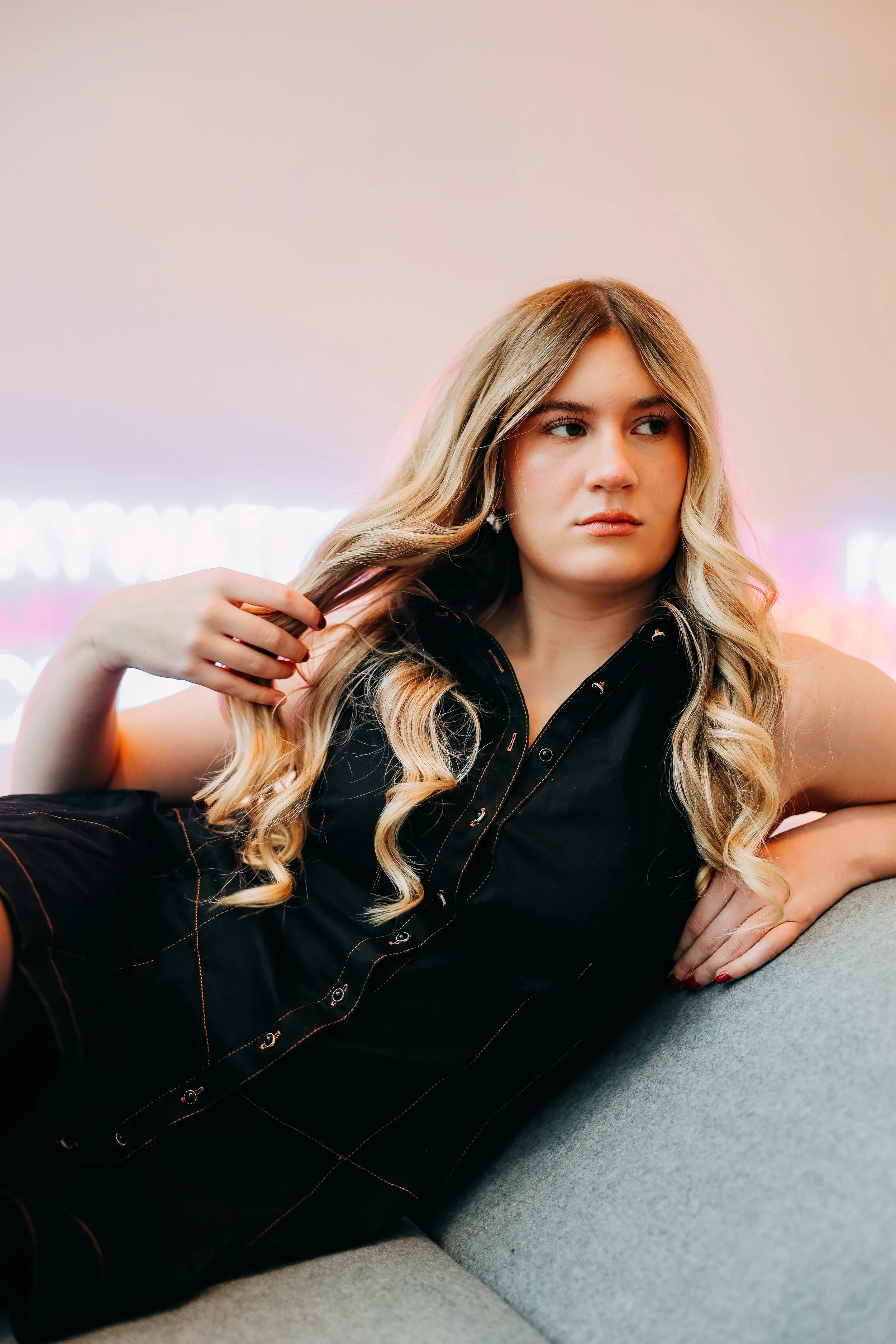A young woman with long blonde curly hair relaxed on a gray couch, wearing a black sleeveless top, with neon lights in the background.