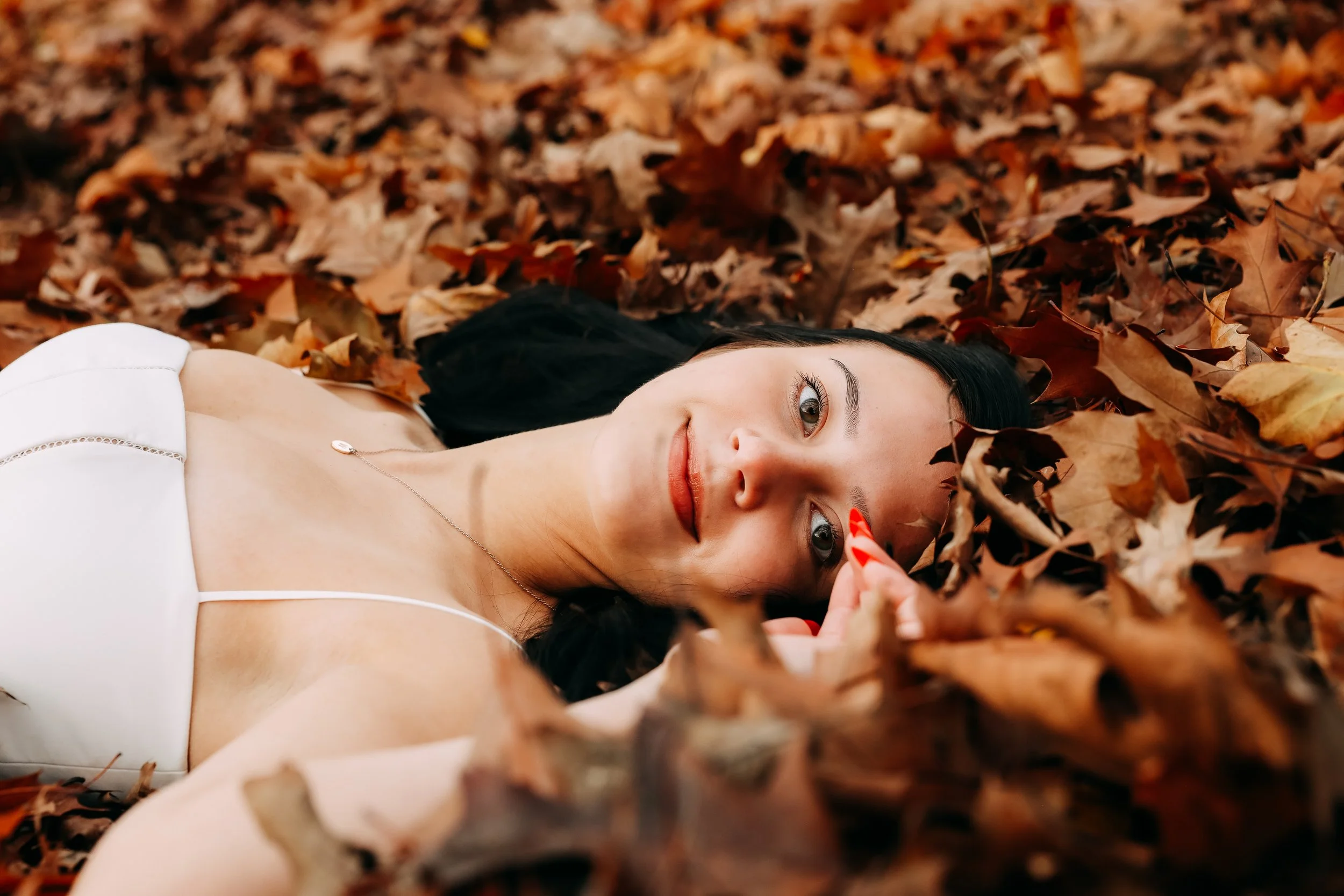 A woman with dark hair and light skin lying on a bed of autumn leaves, looking at the camera, wearing a white top and a delicate necklace.
