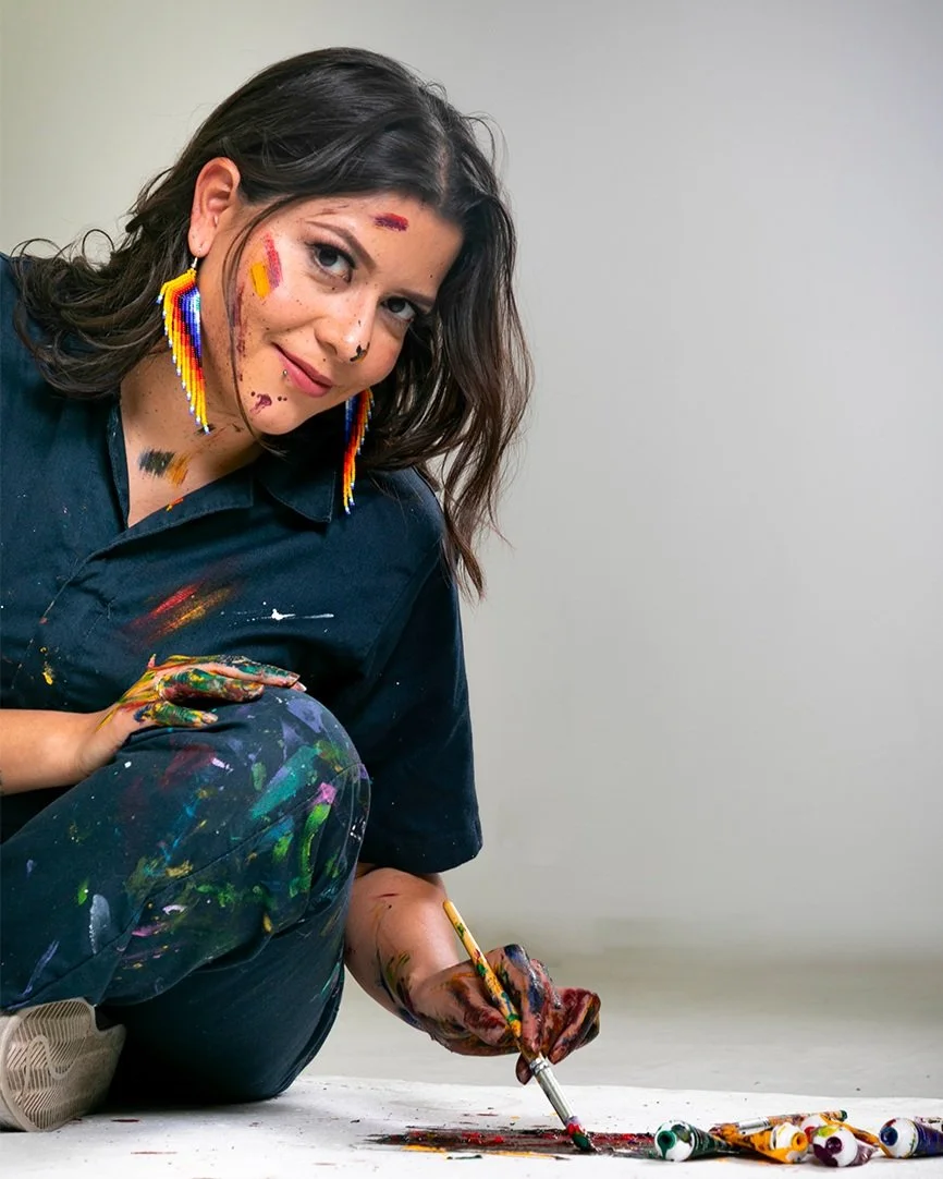 Artist AndreaCataRo sitting on the floor with paint on clothes and face, holding a brush, surrounded by paint tubes.