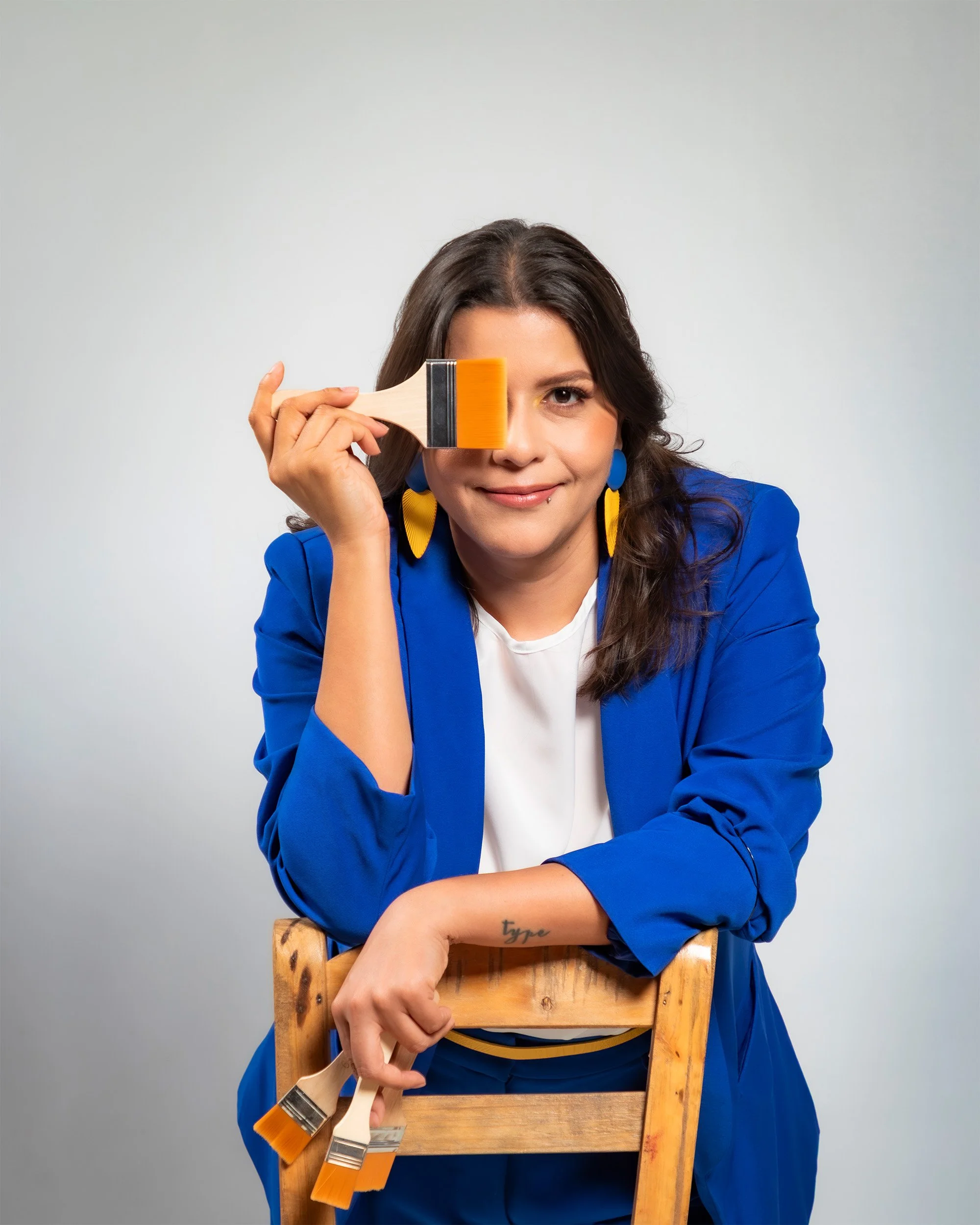 Artist AndreaCataRo is in a blue blazer, leaning on a wooden chair, holding a paintbrush to her right eye, with a playful smile and colourful earrings against a plain background.