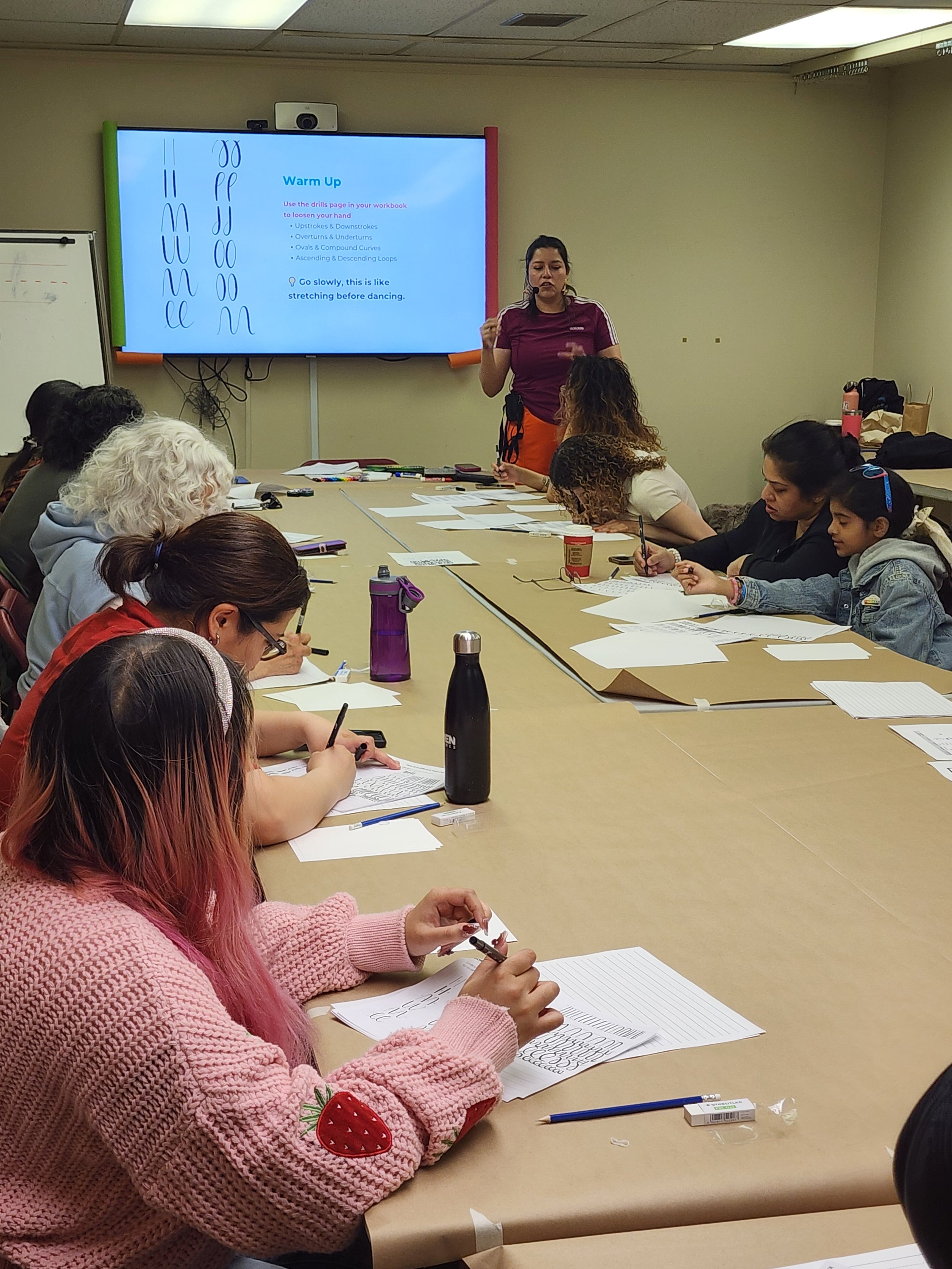A group of children and adults sitting around a conference table, listening to AndreaCataRo, the instructor, lead a calligraphy class. There is a large screen at the front of the room displaying instructions.