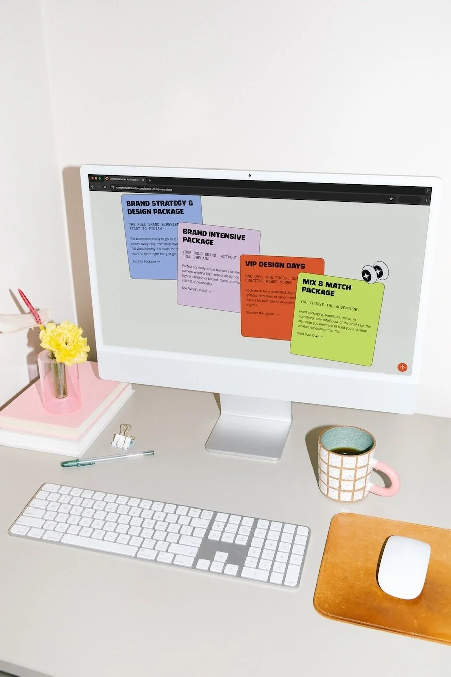 A silver and white desktop computer showing Cheeky Noot Studio's website, sitting next to a pink and white grid pattern mug with black coffee inside, a velvet rusty brown mouse pad, wireless computer mouse and keyboard, stack of notebooks and flowers