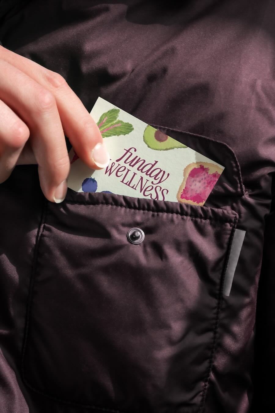 Close-up photo of a woman's hand pulling a business card with Funday Wellness logo, butter yellow background, fruit and vegetable drawings on it out of a purple puffer jacket pocket.