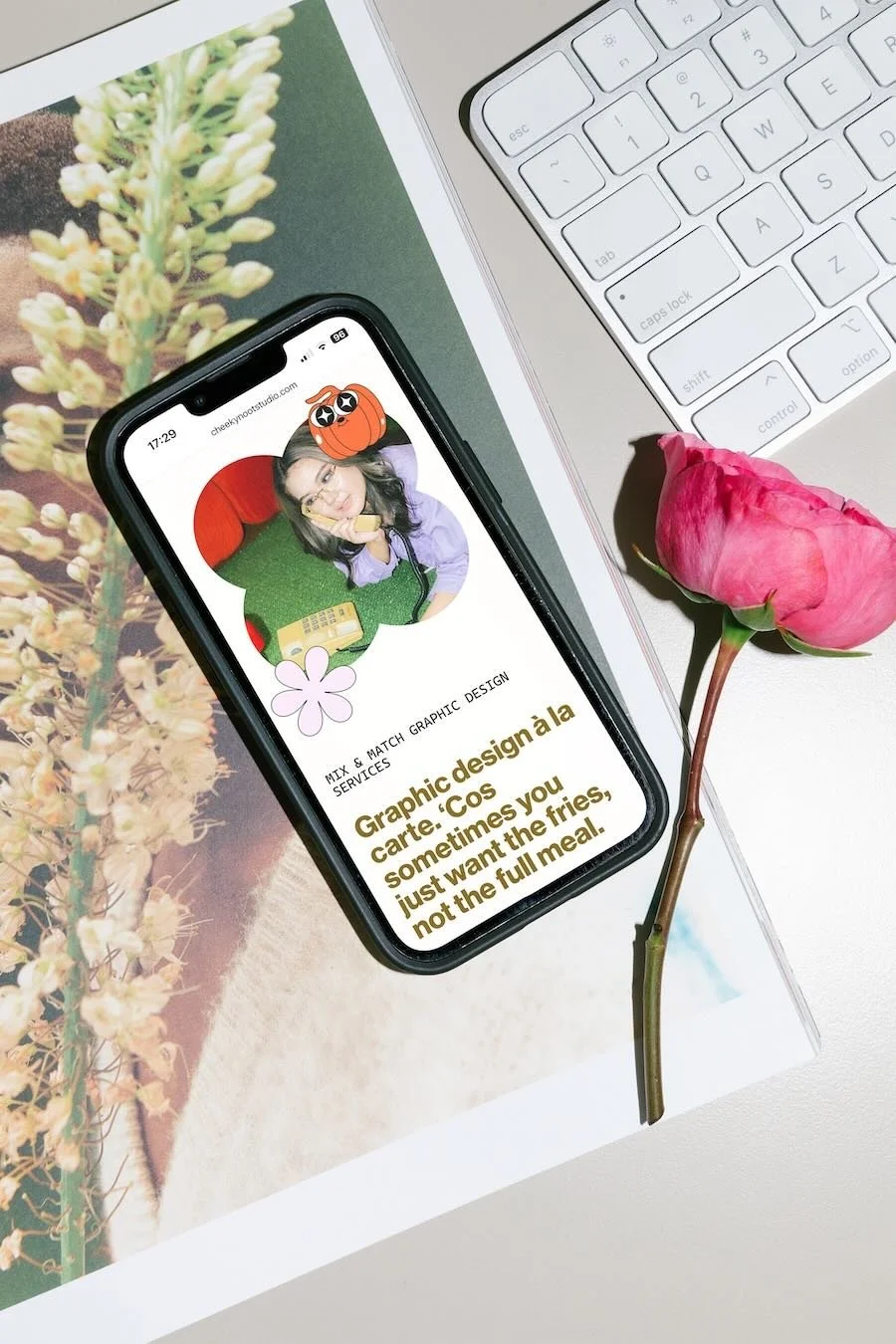 Phone lying on a desk next to an Apple keyboard and a pink flower, showing the mobile Squarespace website for Cheeky Noot.