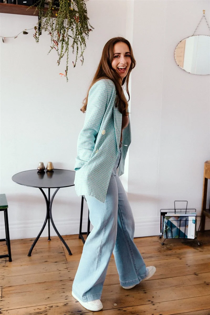 Woman in blue plaid blazer and jeans smiling, walking on wooden floor in a room with plants and a small round table.