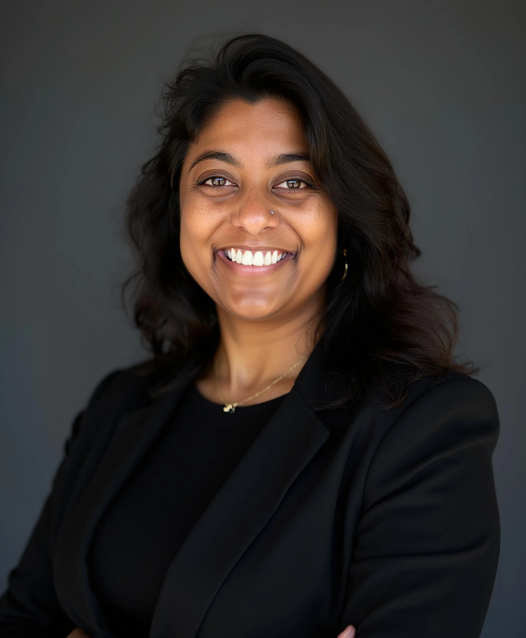 A professional woman with dark hair and brown skin smiling, wearing a black blazer and gold jewelry, against a gray background.