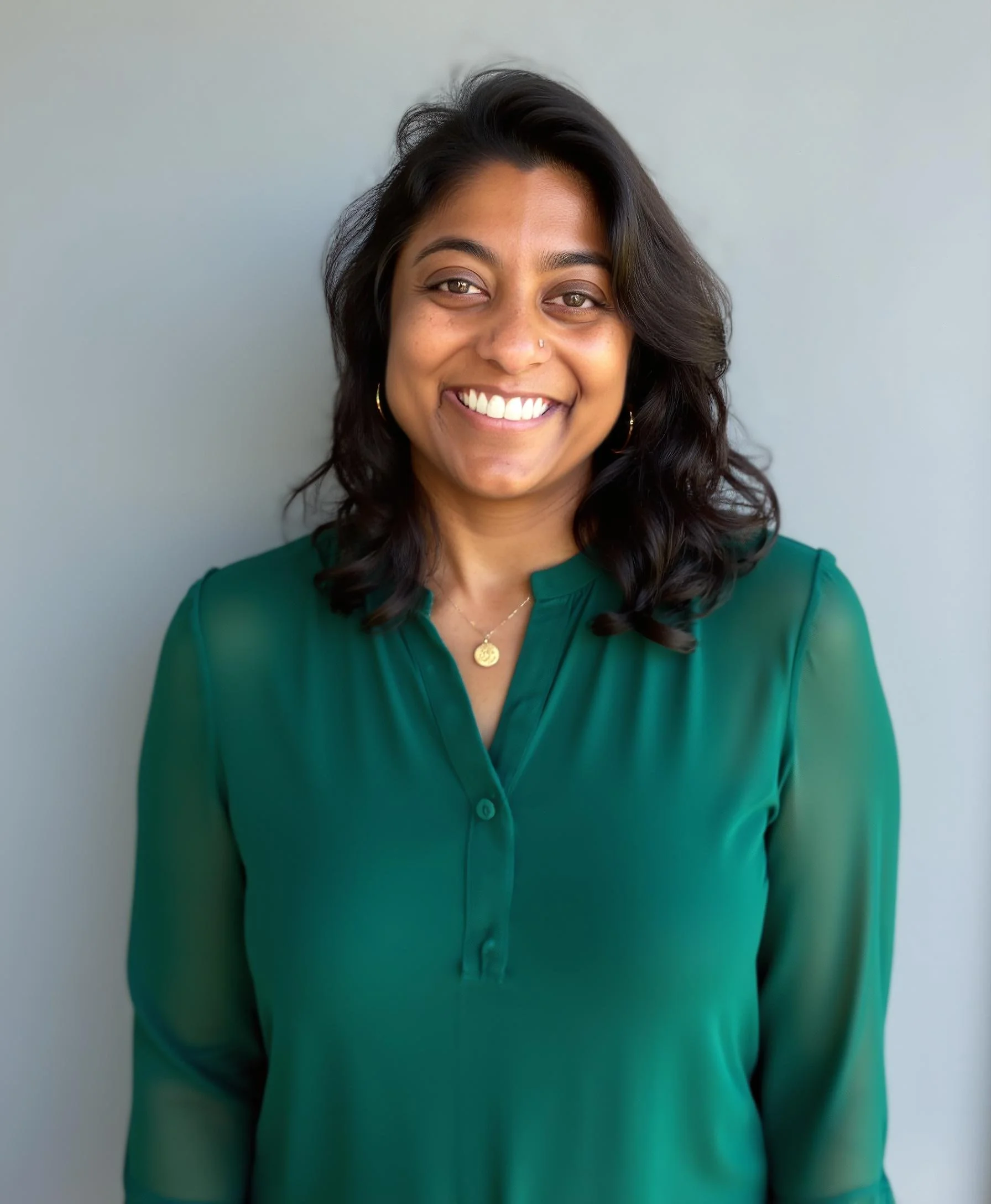 A woman with dark wavy hair wearing a green blouse, smiling at the camera, standing against a gray background.