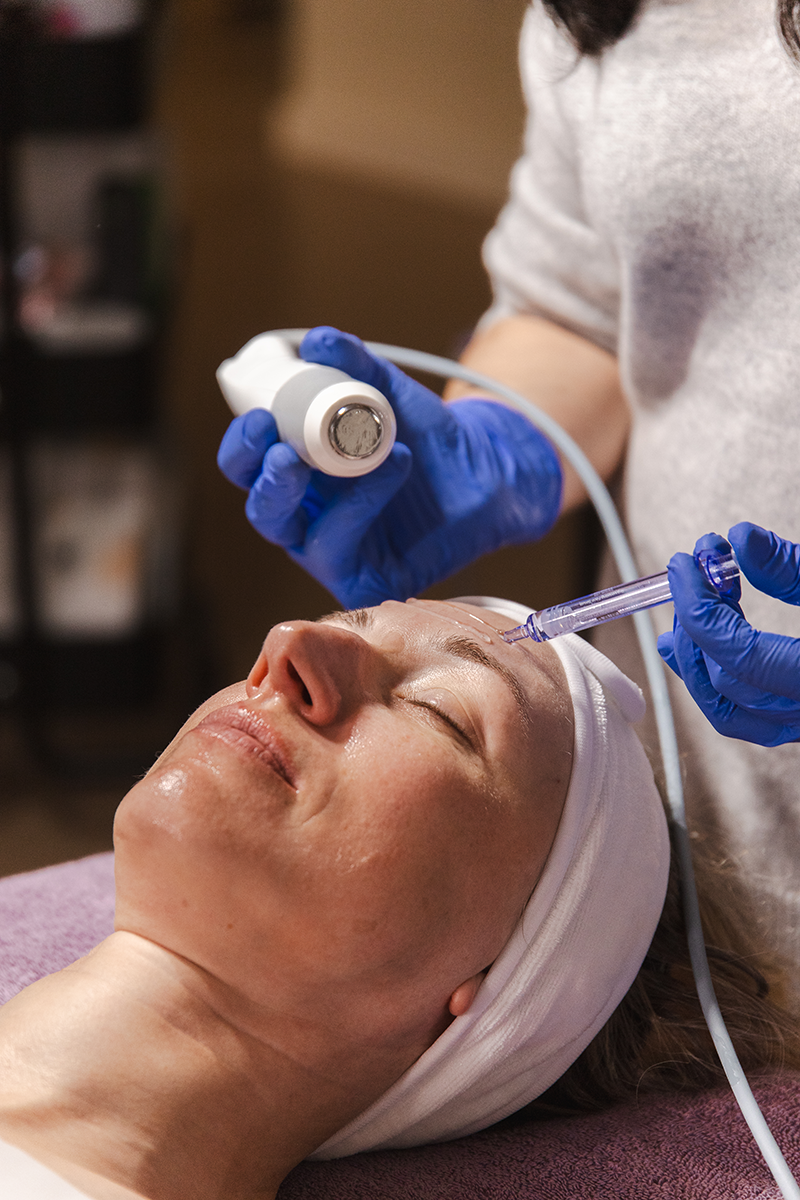 A woman receiving a cosmetic facial treatment with a syringe, while lying down with a headband on, in a skincare clinic setting.