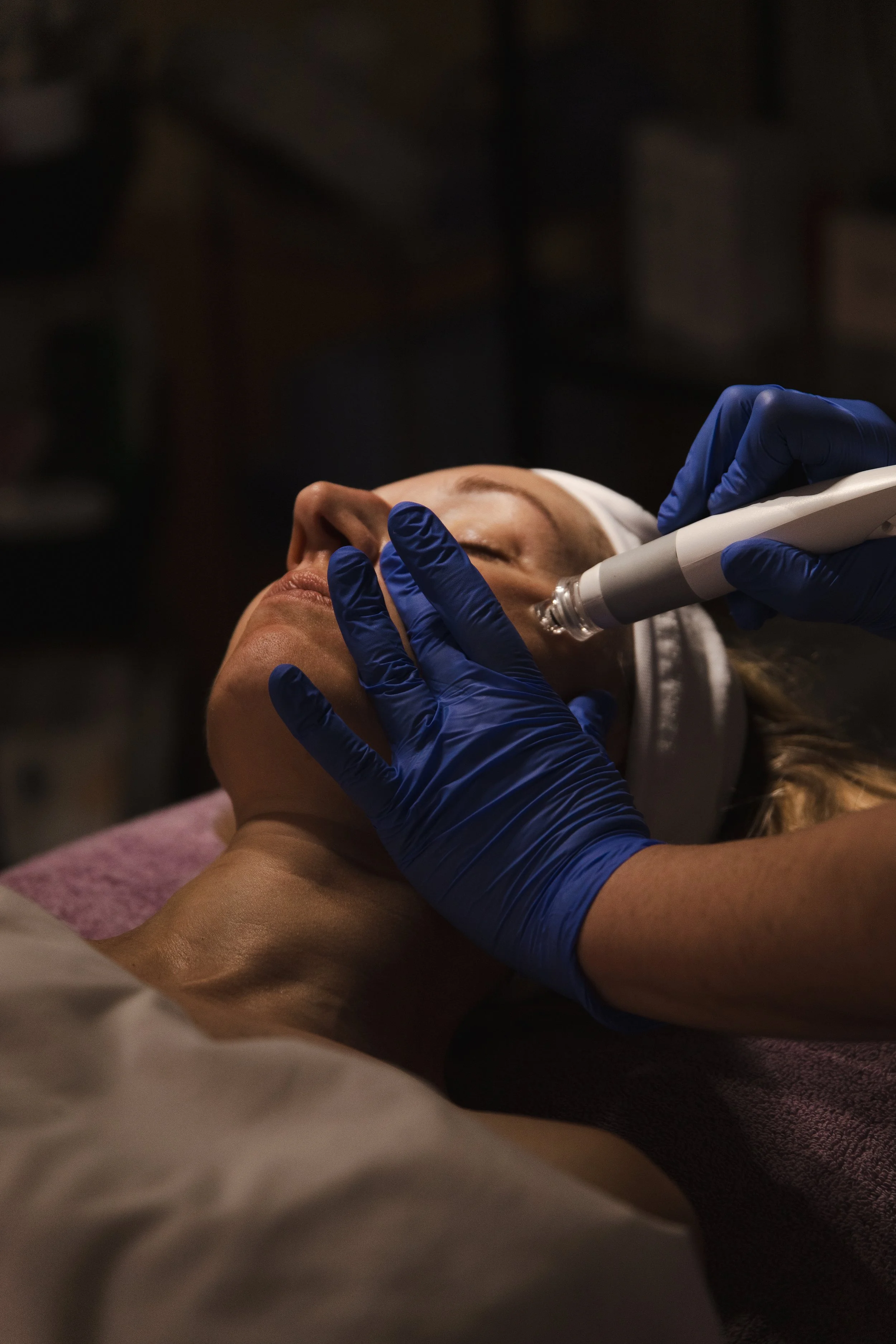 A woman receiving a cosmetic treatment with a handheld device, wearing a headband and lying on a treatment bed, surrounded by a dark background.