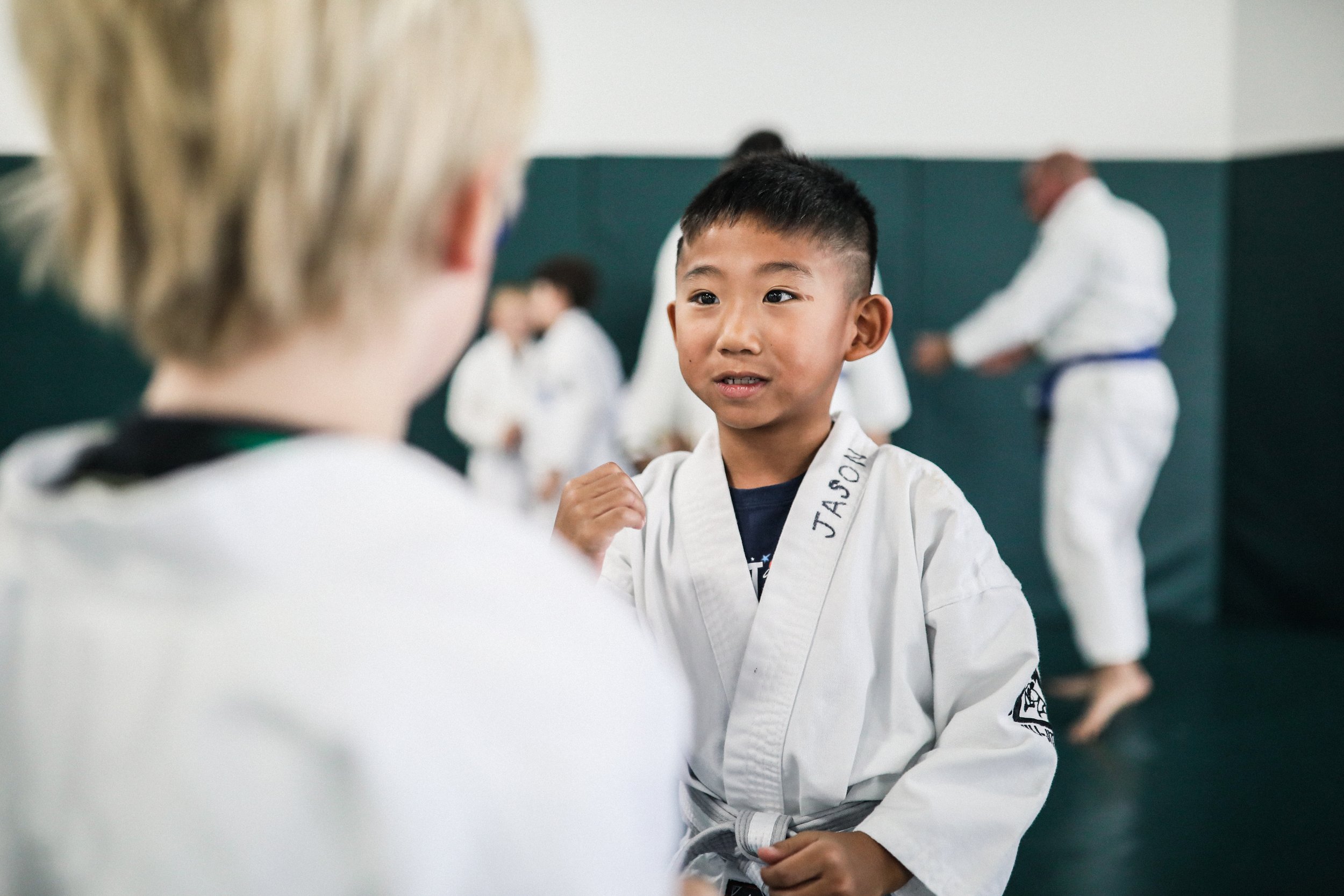 Children practicing martial arts in a dojo, with one boy talking to an instructor or peer, wearing a white gi with 'JASON' on the collar.
