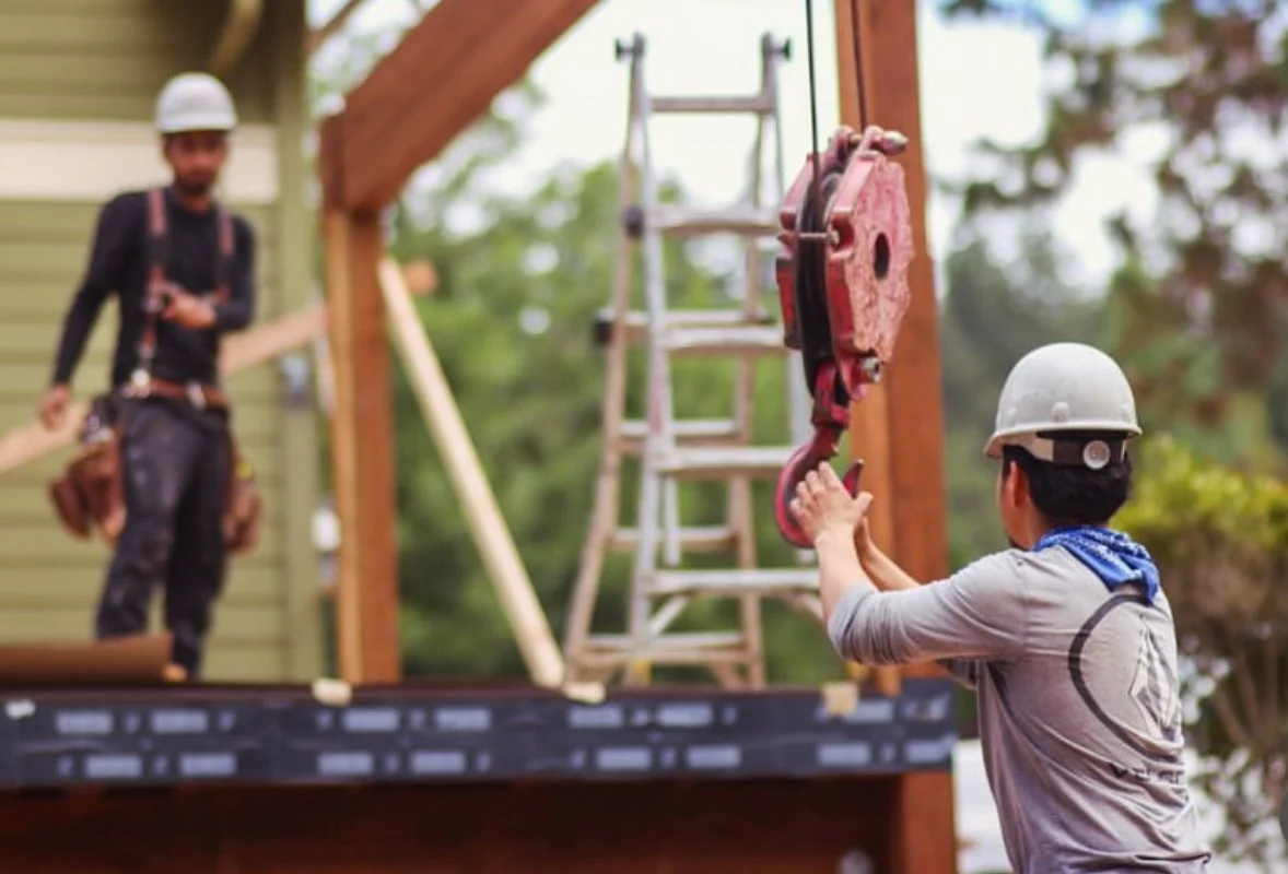 Dos trabajadores en un sitio de construcción, uno colocando una pieza de equipo y otro sosteniendo una cuerda, rodeados de estructura de madera y herramientas.