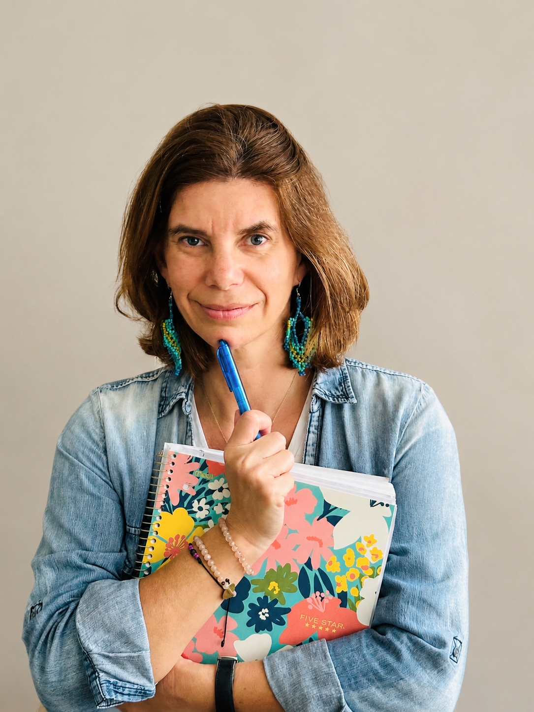 Woman with brown hair holding a floral notebook and a blue pen, wearing earrings and bracelets, standing against a plain background.