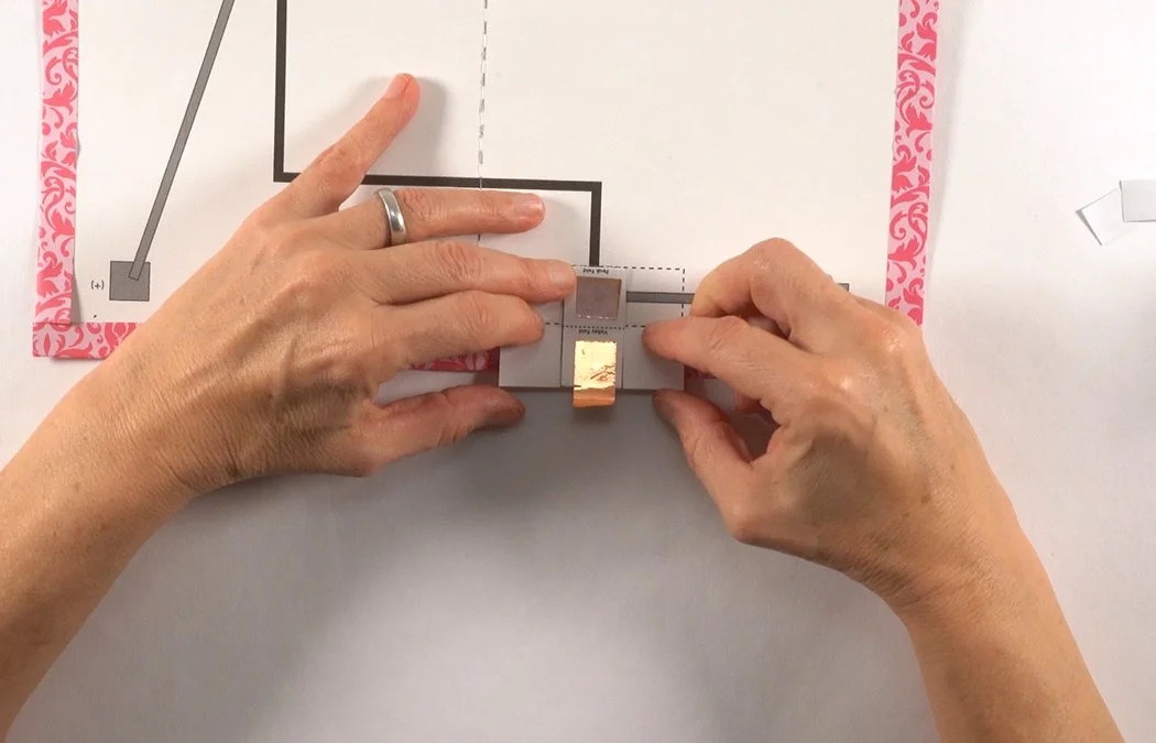 Place it directly over the grey (+) rectangle inside the battery box. Make sure neither copper gets too close to the dashed fold line.