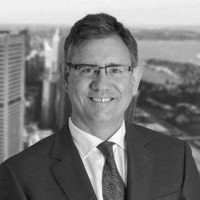 Black and white photo of a smiling man in a suit standing on wooden stairs indoors, with a modern architectural background.