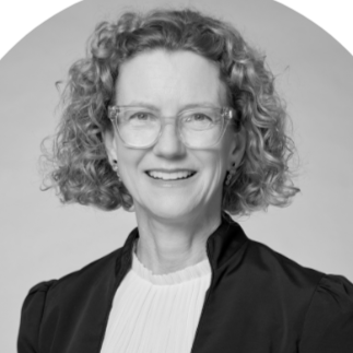 Black and white photo of a smiling woman with curly hair, wearing glasses, a black jacket, and a white blouse.