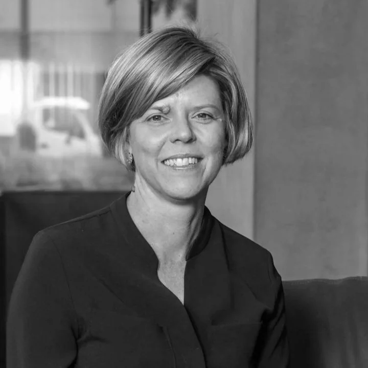 A smiling woman with short, blonde hair, wearing a dark blazer, sitting indoors against a blurred background.