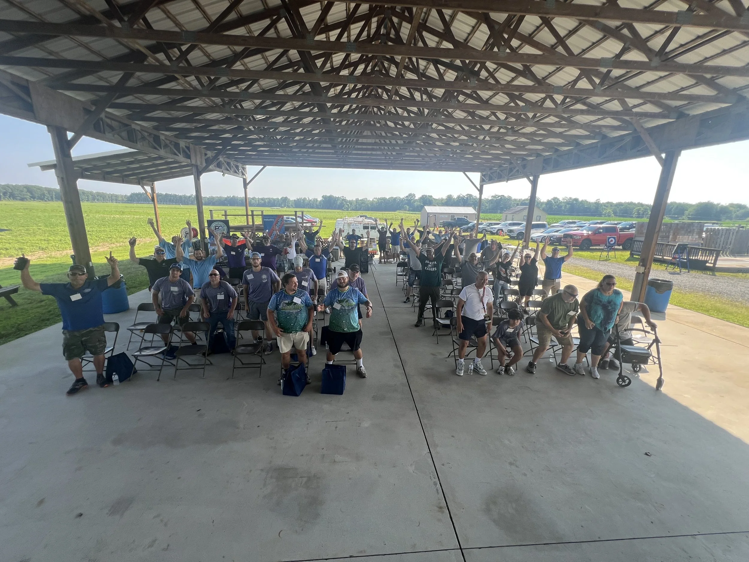 People sitting and standing under an open-sided shelter at an outdoor event with a field, parking lot, and clear sky in the background.