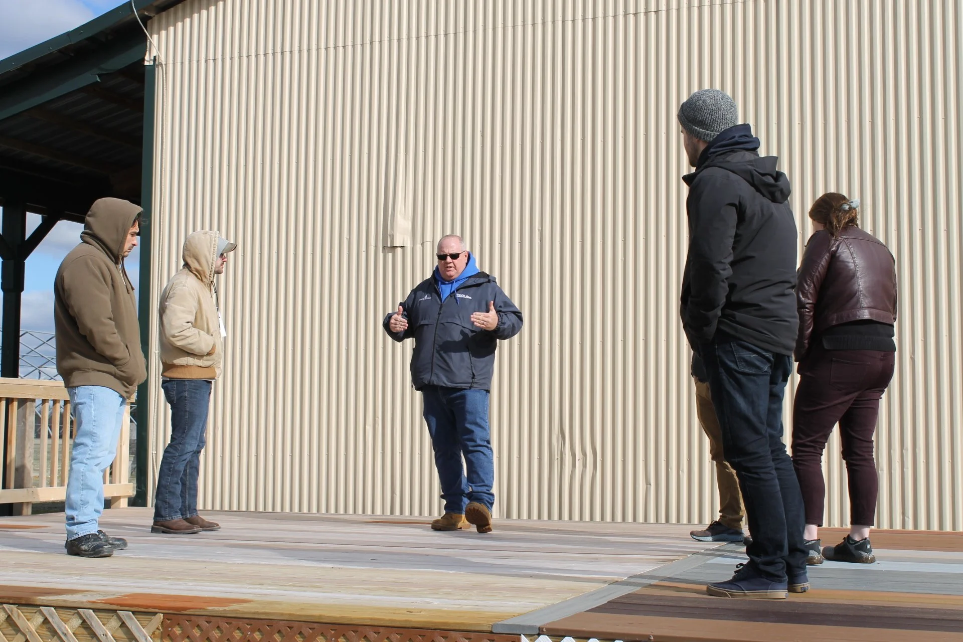 A group of people standing outdoors in front of a large beige corrugated metal building, listening to a man in sunglasses and a dark jacket who appears to be giving a speech or instructions.
