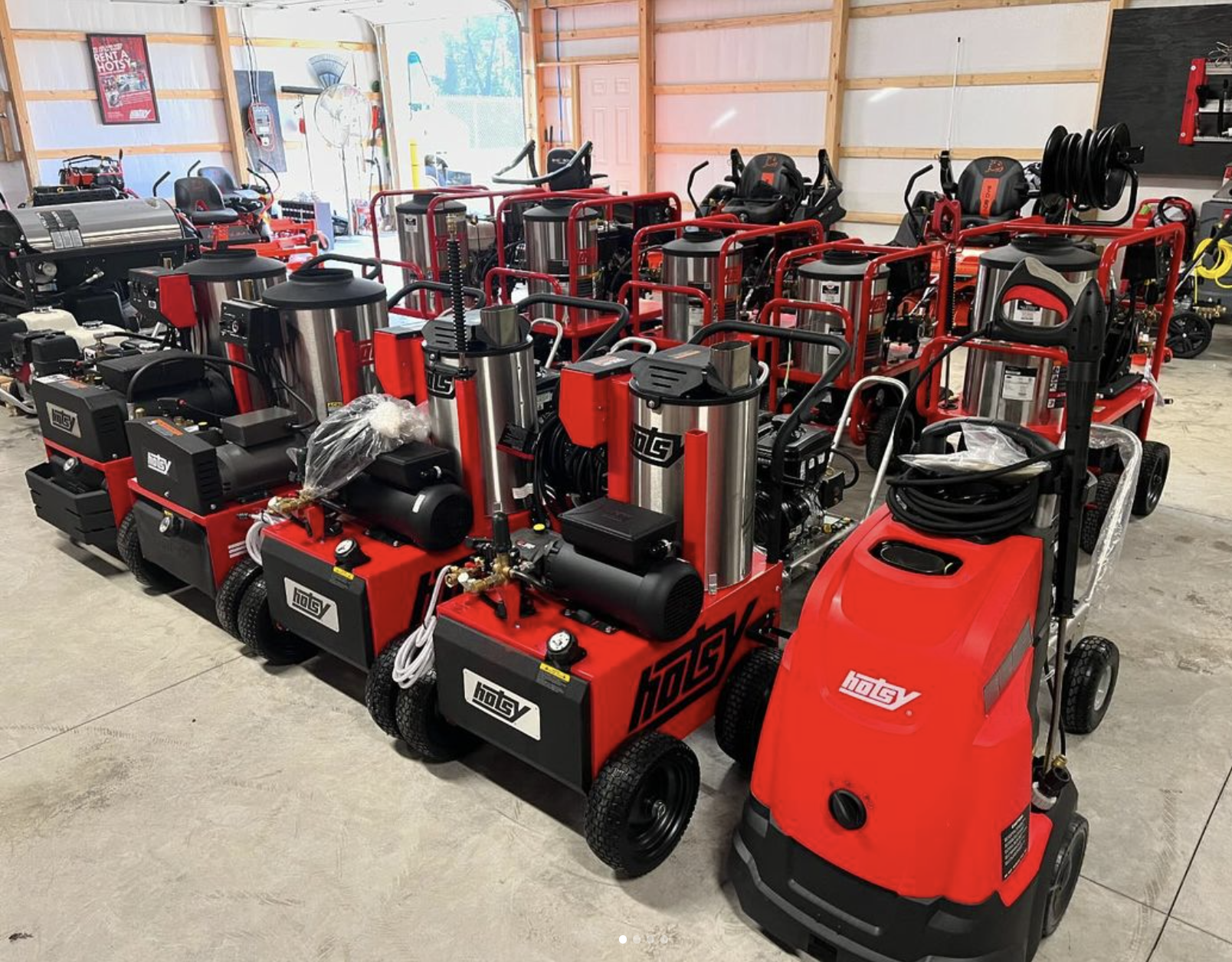 A collection of red industrial pressure washers with black and silver components, arranged in rows inside a storage or workshop space with wooden framing and a concrete floor.