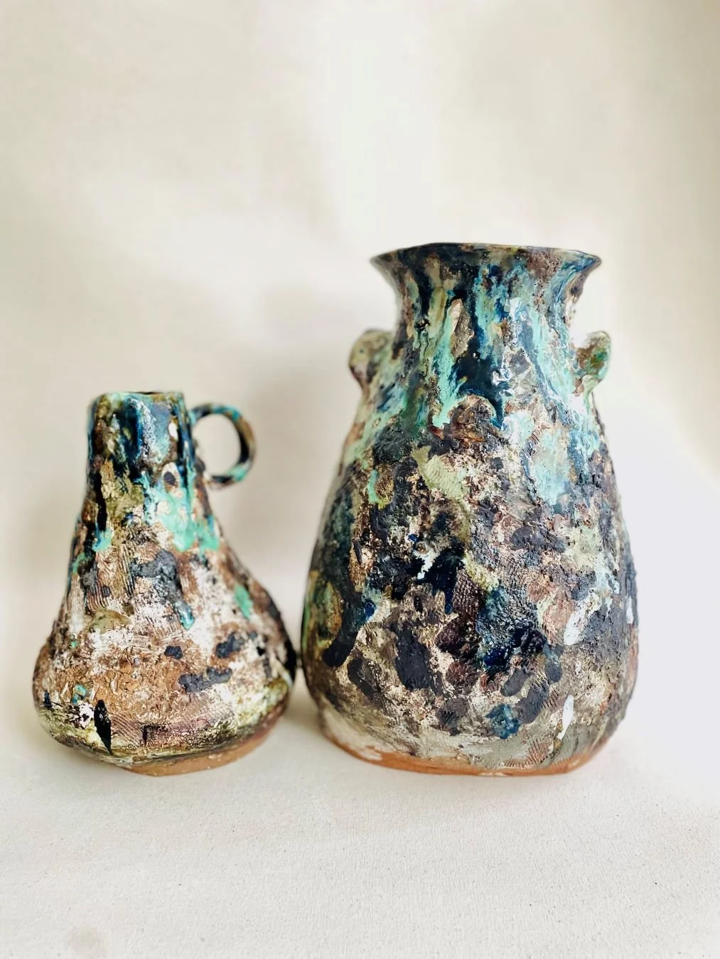 Two abstract, textured ceramic vases with a multicolored, mottled glaze finish, one larger and one smaller, set against a plain light-colored background.