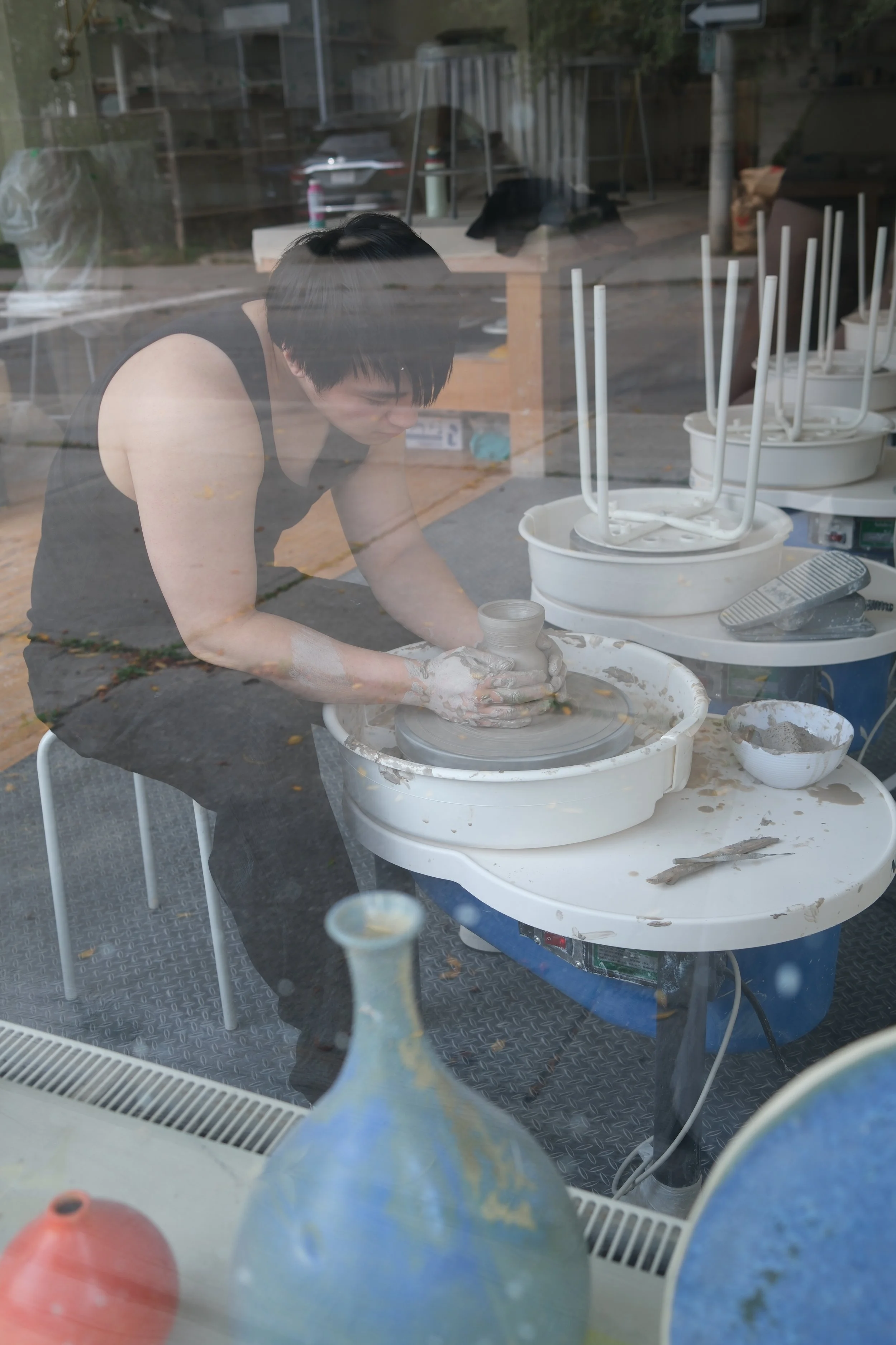 Reflected image of a person with short dark hair working with pottery on a pottery wheel inside a studio. Outside the studio window, there is a fenced yard with a bear-like figure on a bench, and stacked chairs on tables near the window.