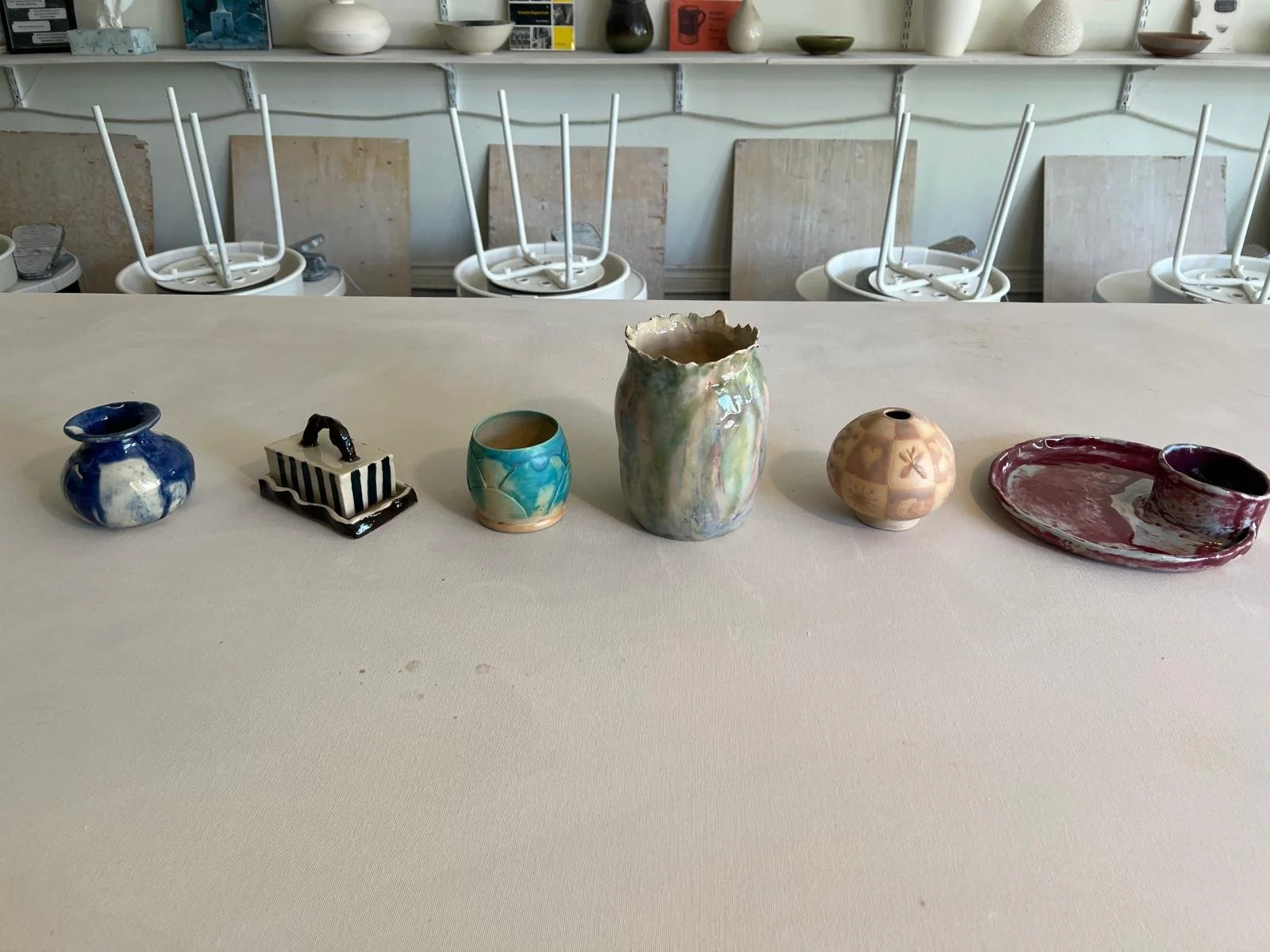 A row of seven small, colorful, handcrafted pottery pieces on a white table with a workshop or studio background.