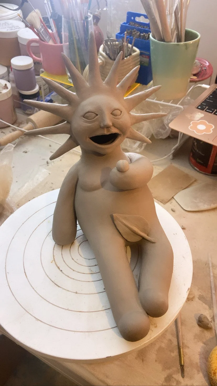 A ceramic sculpture of a humanoid figure with a sun-like head featuring elongated spikes. The figure is seated with one arm resting on its lap, smiling widely with a large open mouth, and appears to be pregnant, with one hand on its belly. The sculpture is on a pottery wheel in a workspace with pottery tools and supplies in the background.