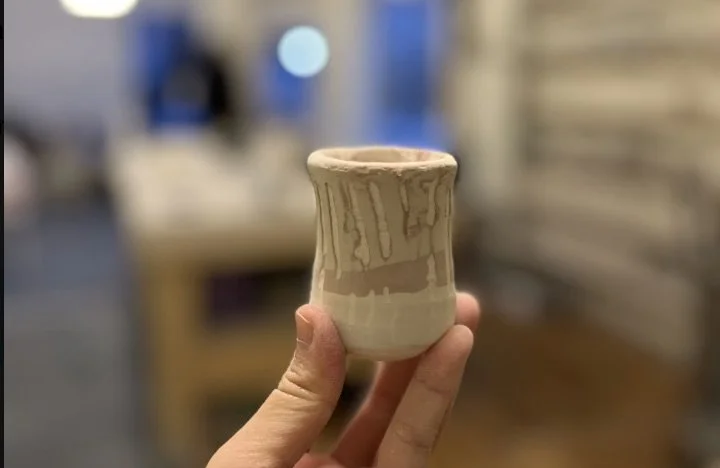 A hand holding a small, beige ceramic cup with a textured, dripping glaze design, against a blurred indoor background.