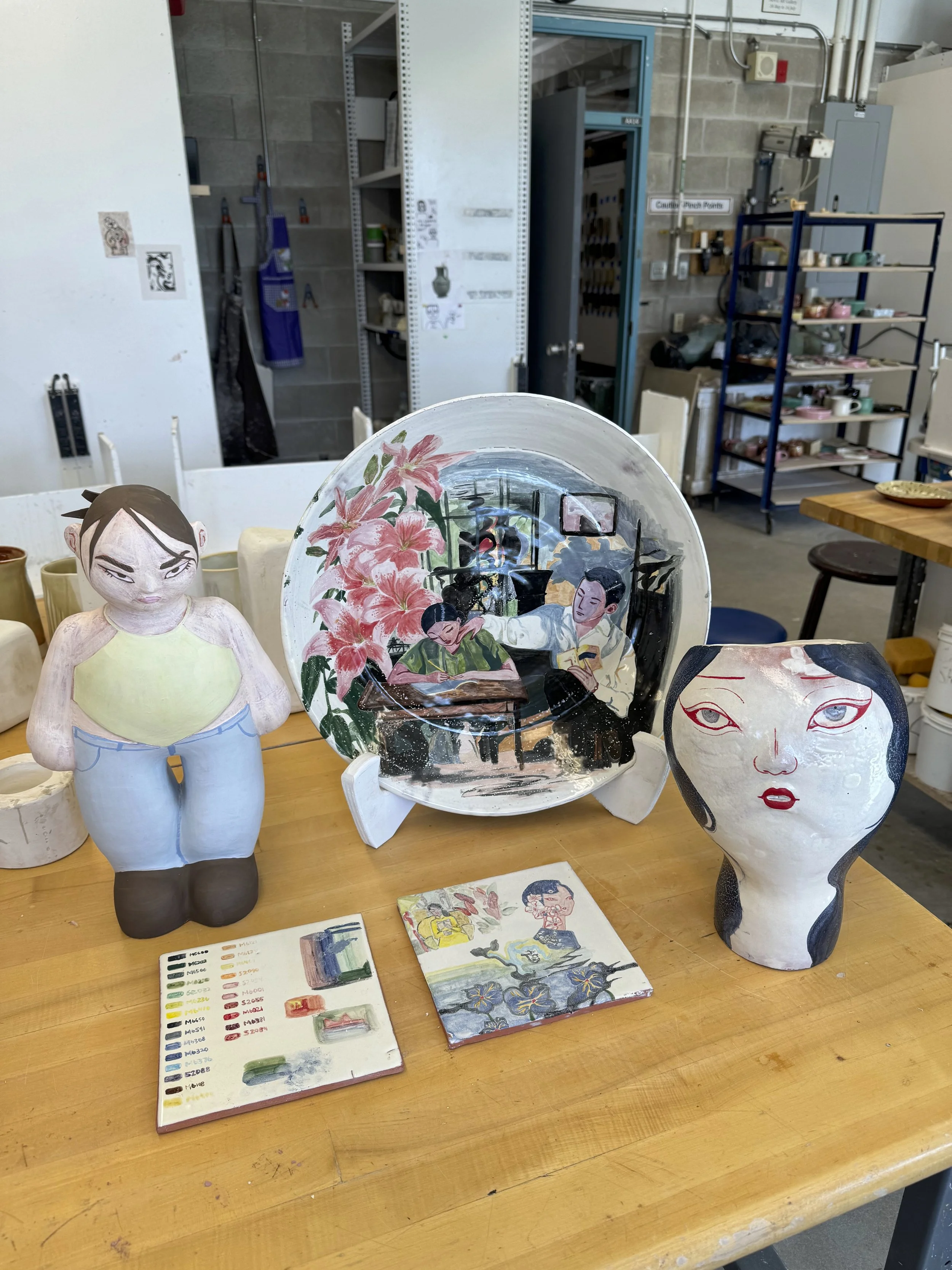 Artistic sculptures of women and ornate painted plates are displayed on a wooden table in an art studio or classroom. In the background, there is a metal shelf with art supplies and a cluttered workspace.